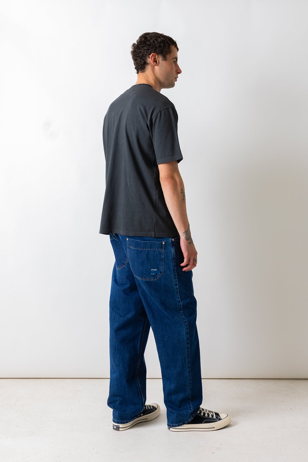 A man wearing a dark charcoal gray t-shirt and blue denim jeans, shown from a side-back profile against a plain white studio background.