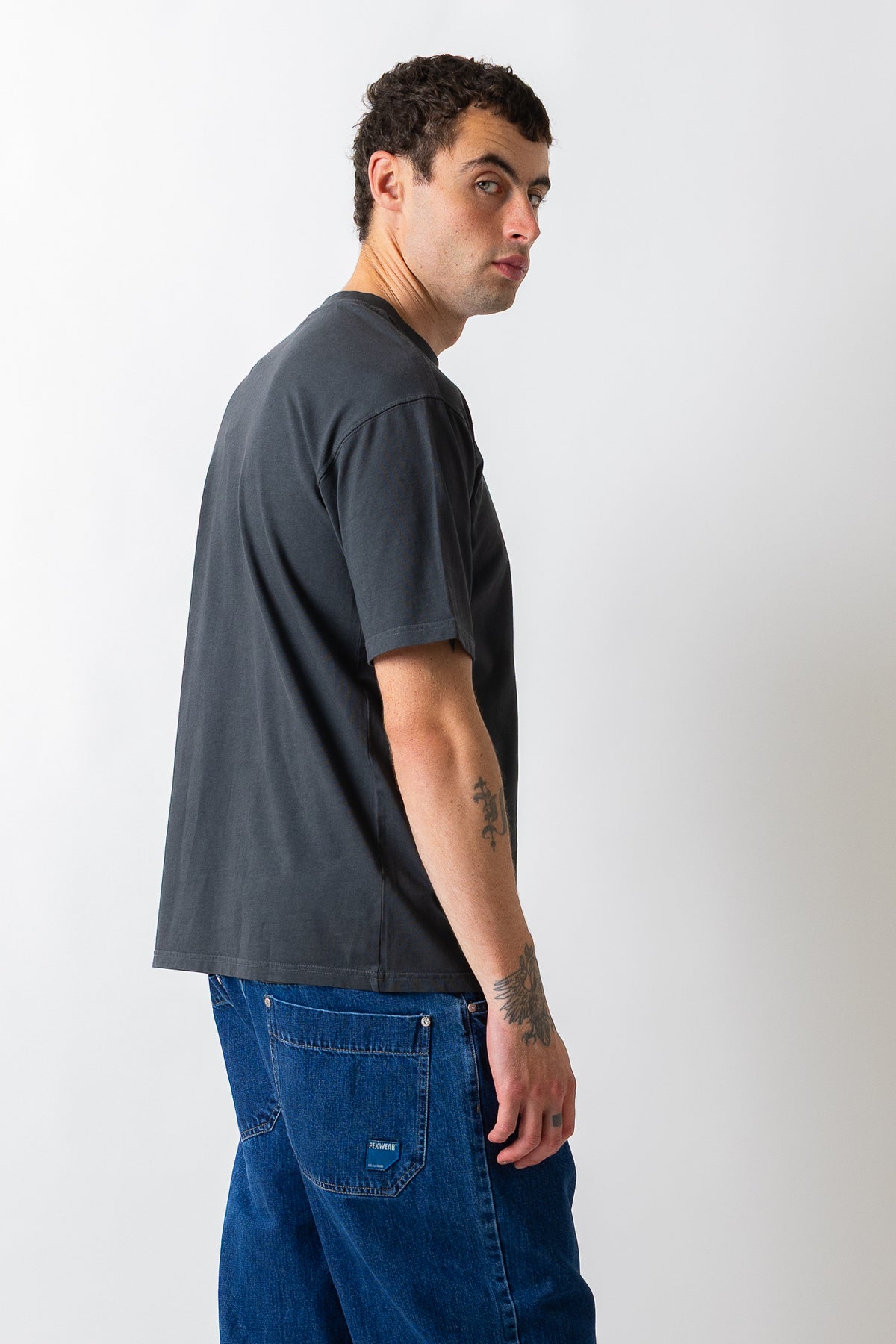 A man wearing a dark charcoal gray t-shirt and blue jeans, shown from a side-back profile against a plain light gray studio background.
