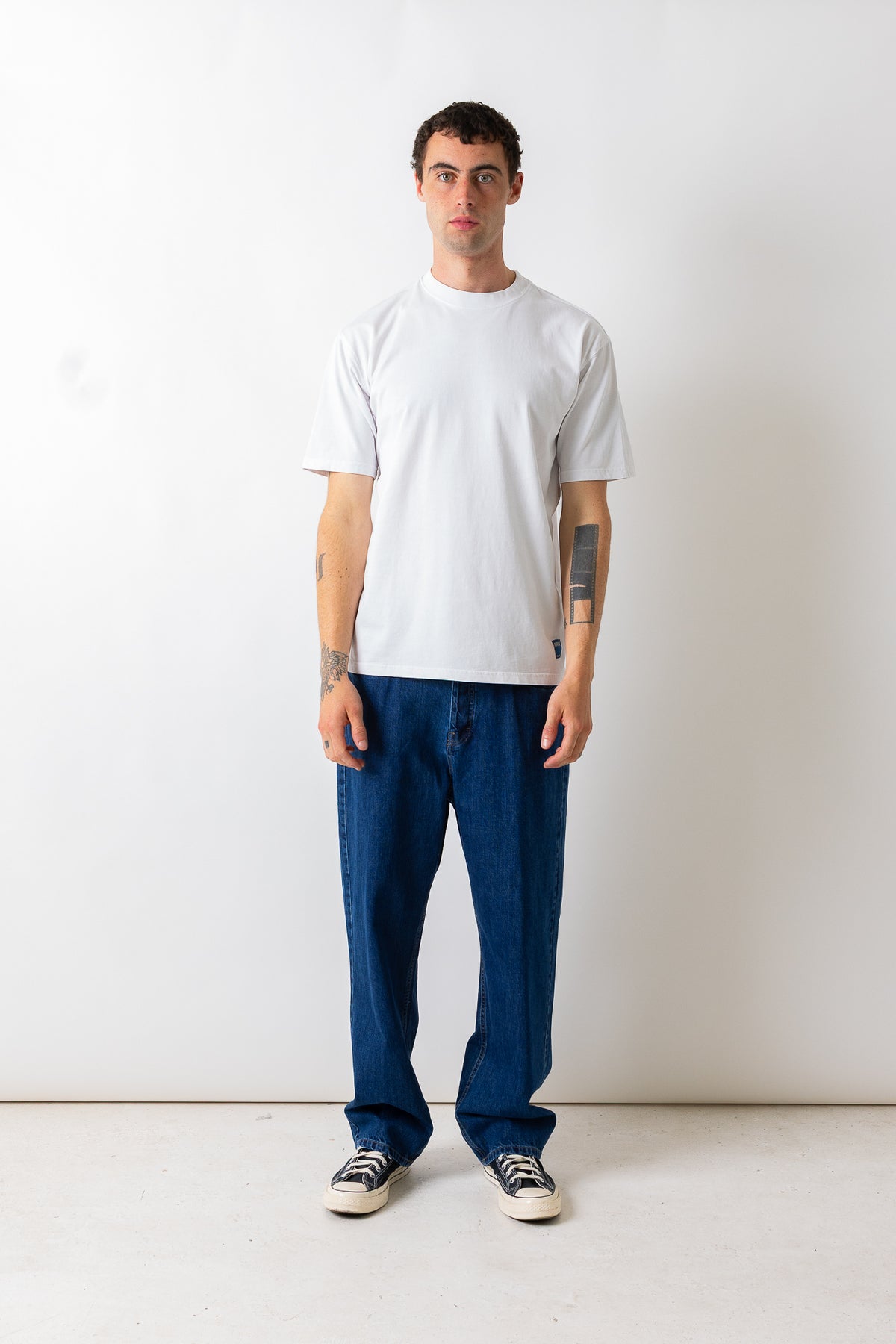 A man stands against a plain white wall wearing a plain white short-sleeve t-shirt, loose dark blue jeans, and black-and-white sneakers.