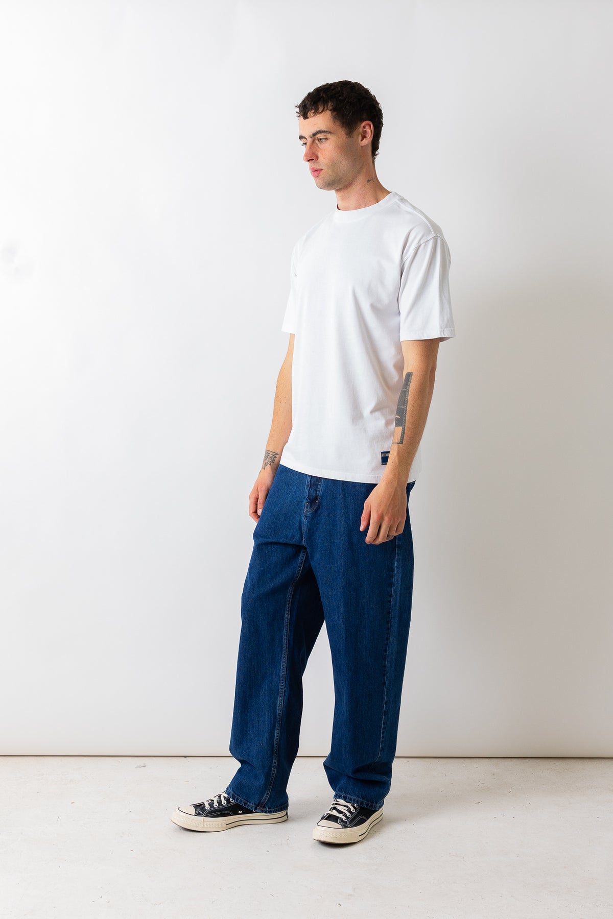 A man wearing a white crew-neck t-shirt and loose-fit dark blue jeans, standing in profile against a plain white studio wall.