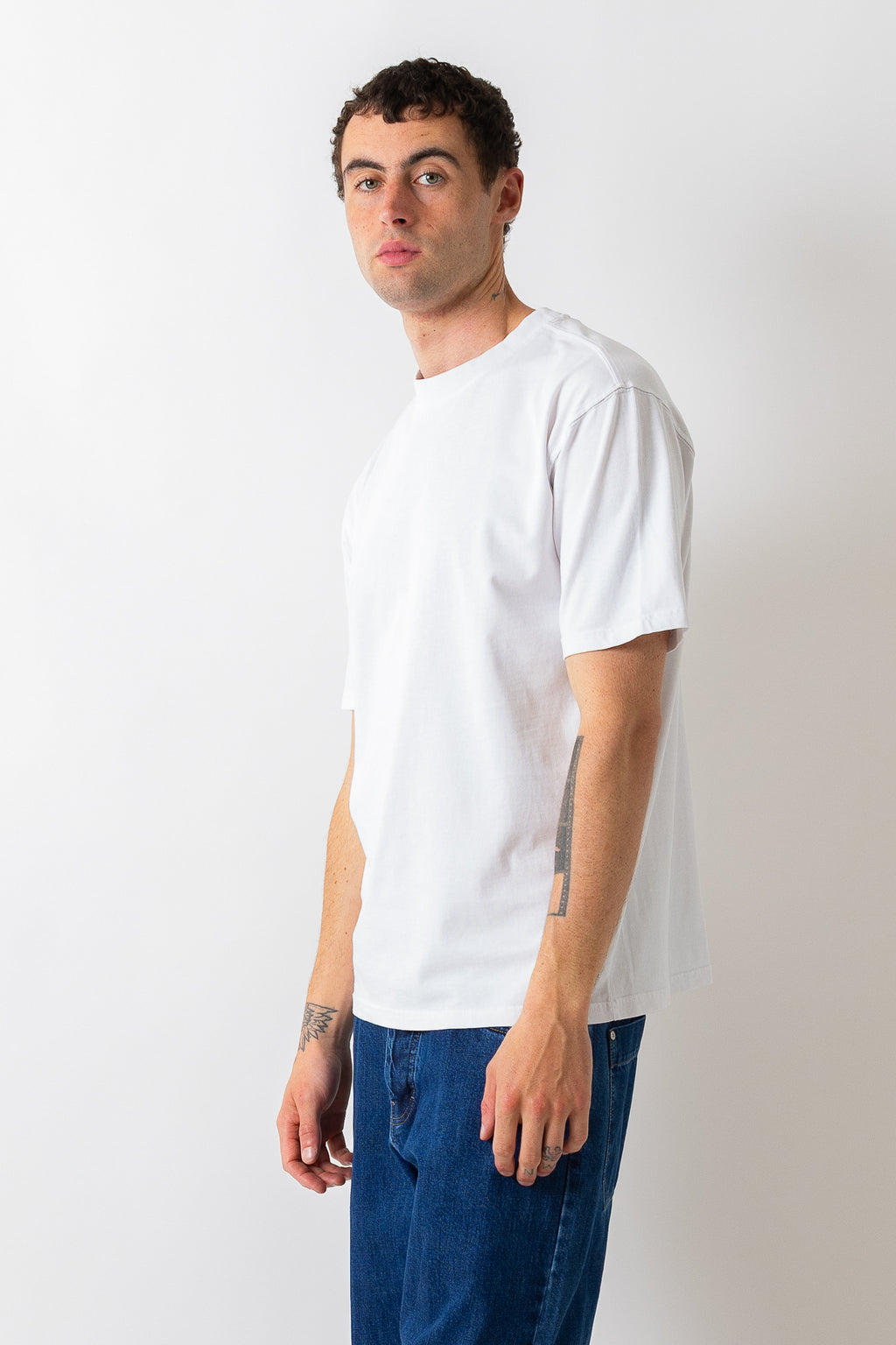 A man wearing a plain white short-sleeve crewneck t-shirt and blue jeans, standing against a light gray studio background.