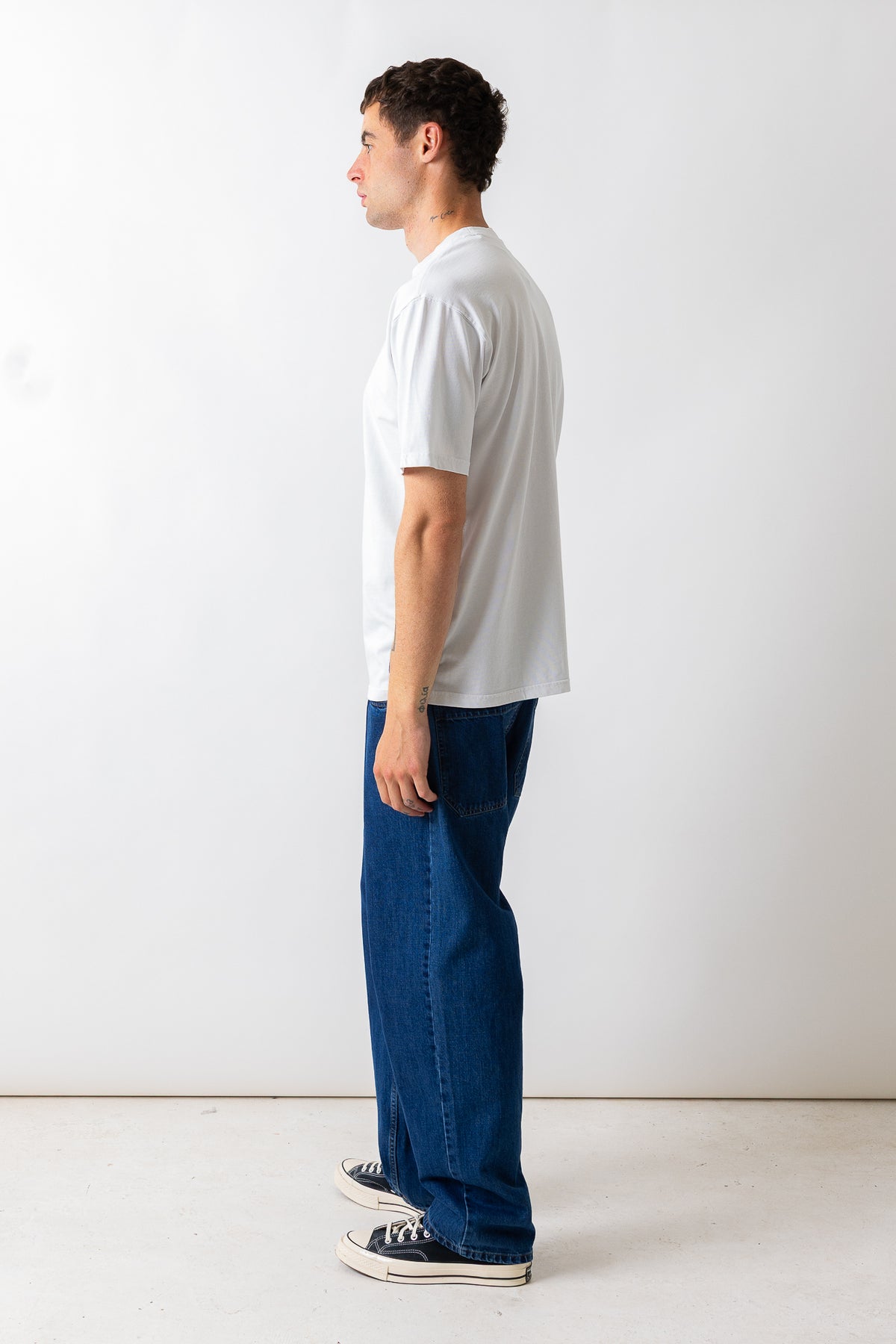 A man standing in profile wearing a white short-sleeve crewneck t-shirt, loose-fit dark blue jeans, and black canvas sneakers against a plain white studio background.