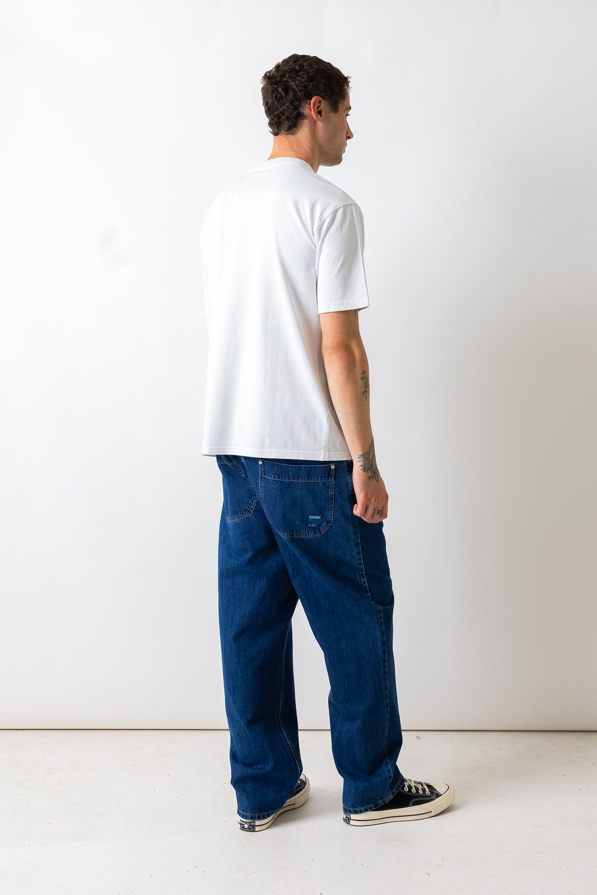 A man wearing a white t-shirt and dark blue relaxed-fit jeans, shown from a back-three-quarter view against a plain white studio background.
