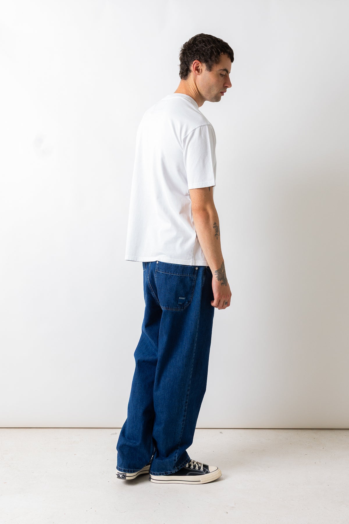 A man wearing a white crewneck t-shirt and loose-fit dark blue denim jeans, standing in profile against a plain white studio wall.