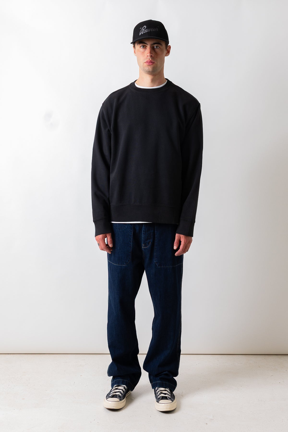 A man stands facing forward in a studio wearing a black crewneck sweatshirt, dark blue jeans, black and white sneakers, and a black baseball cap.