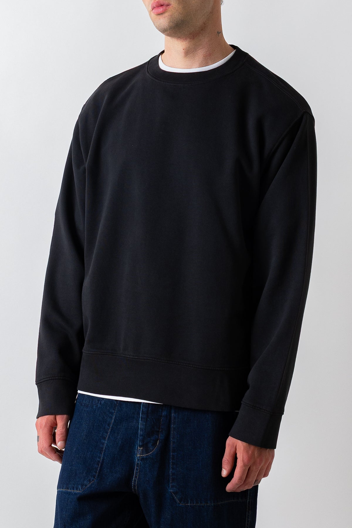 A man wearing a plain black crewneck sweatshirt over a white undershirt and dark blue jeans, photographed against a light gray studio background.