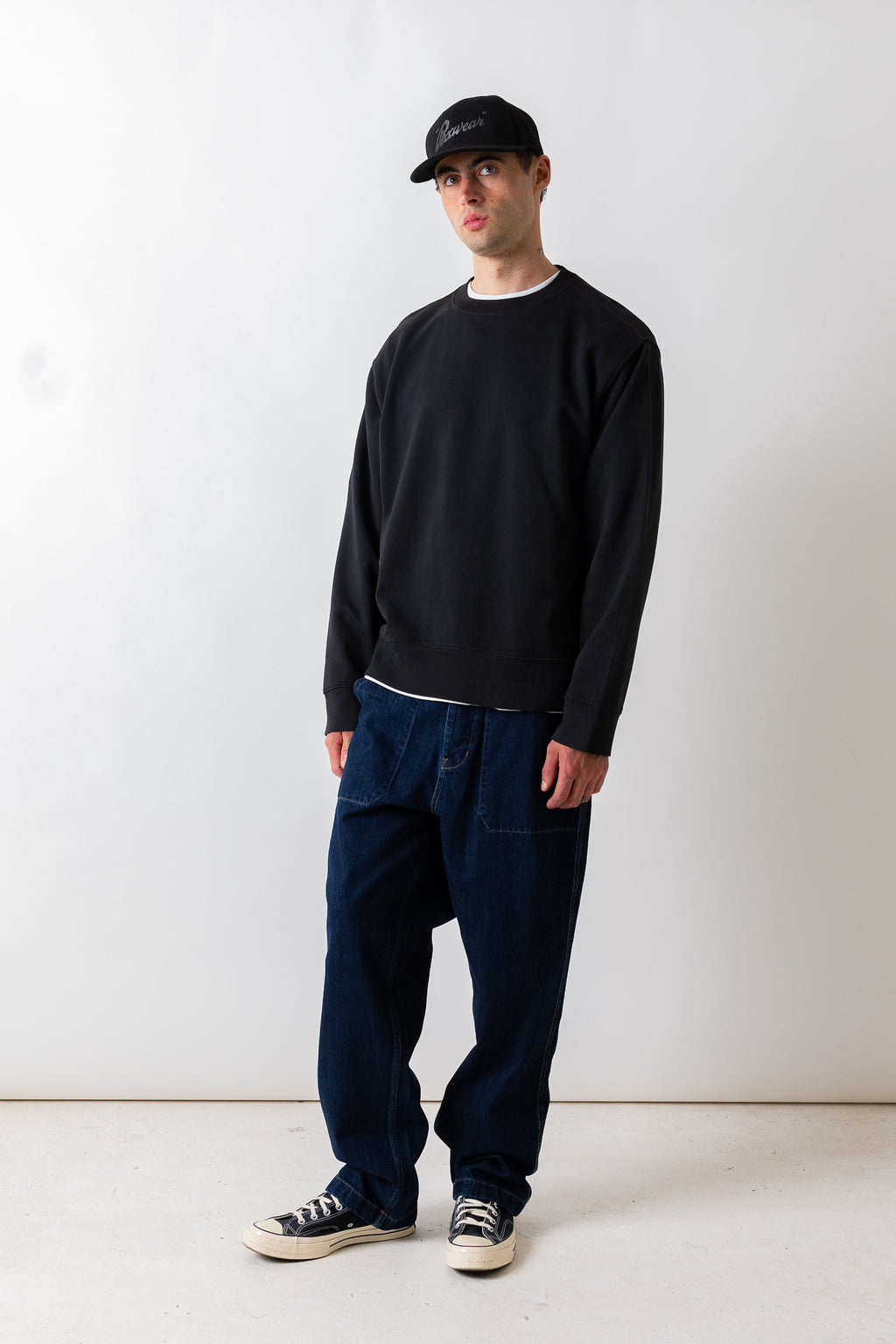 A man wearing a black crewneck sweatshirt, dark blue relaxed-fit jeans, black and white sneakers, and a black baseball cap, standing against a plain white studio background.