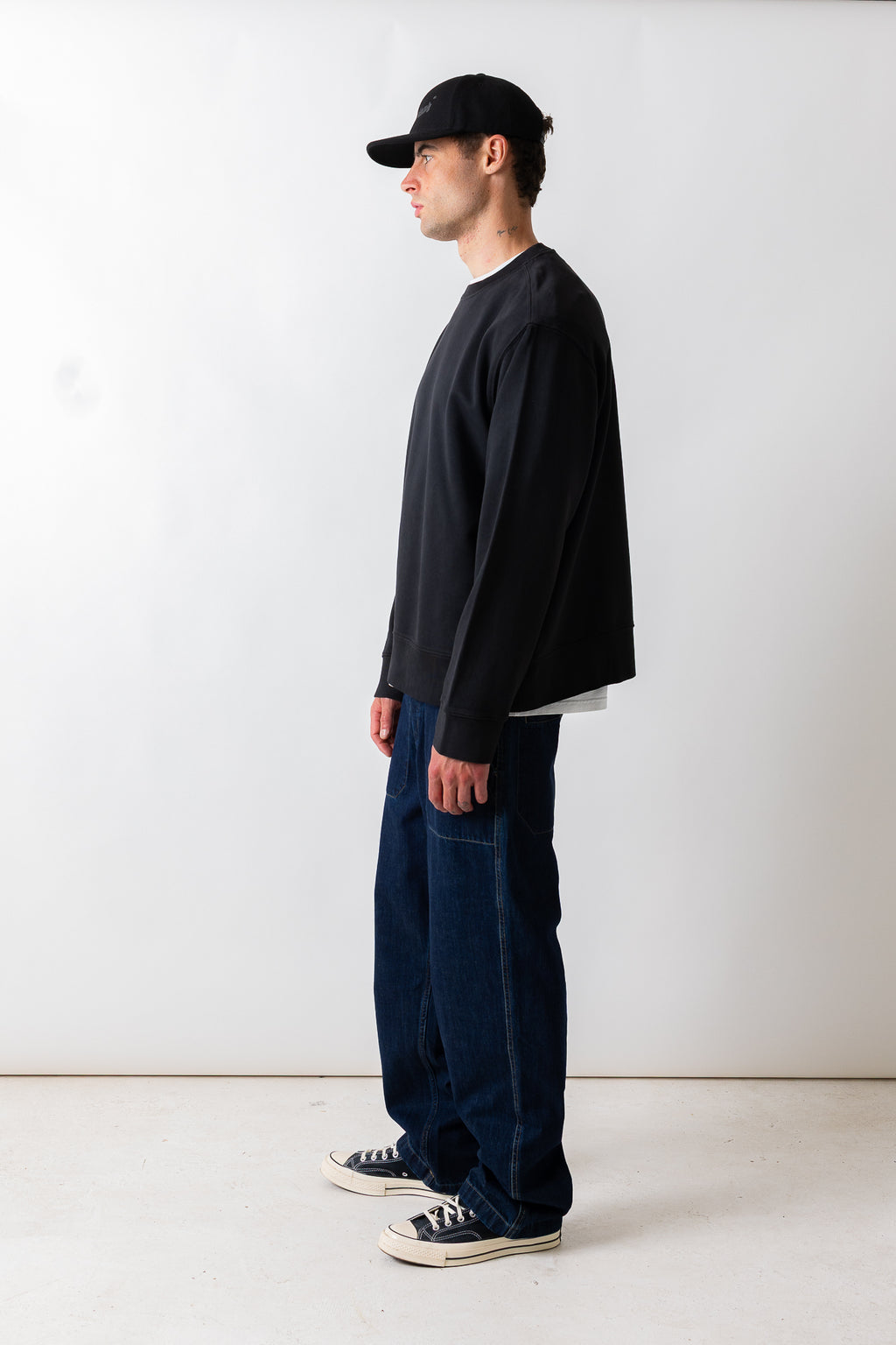 A man in profile wearing a black long-sleeve crewneck sweatshirt, dark blue baggy jeans, a black baseball cap, and black canvas sneakers, standing against a plain white studio background.