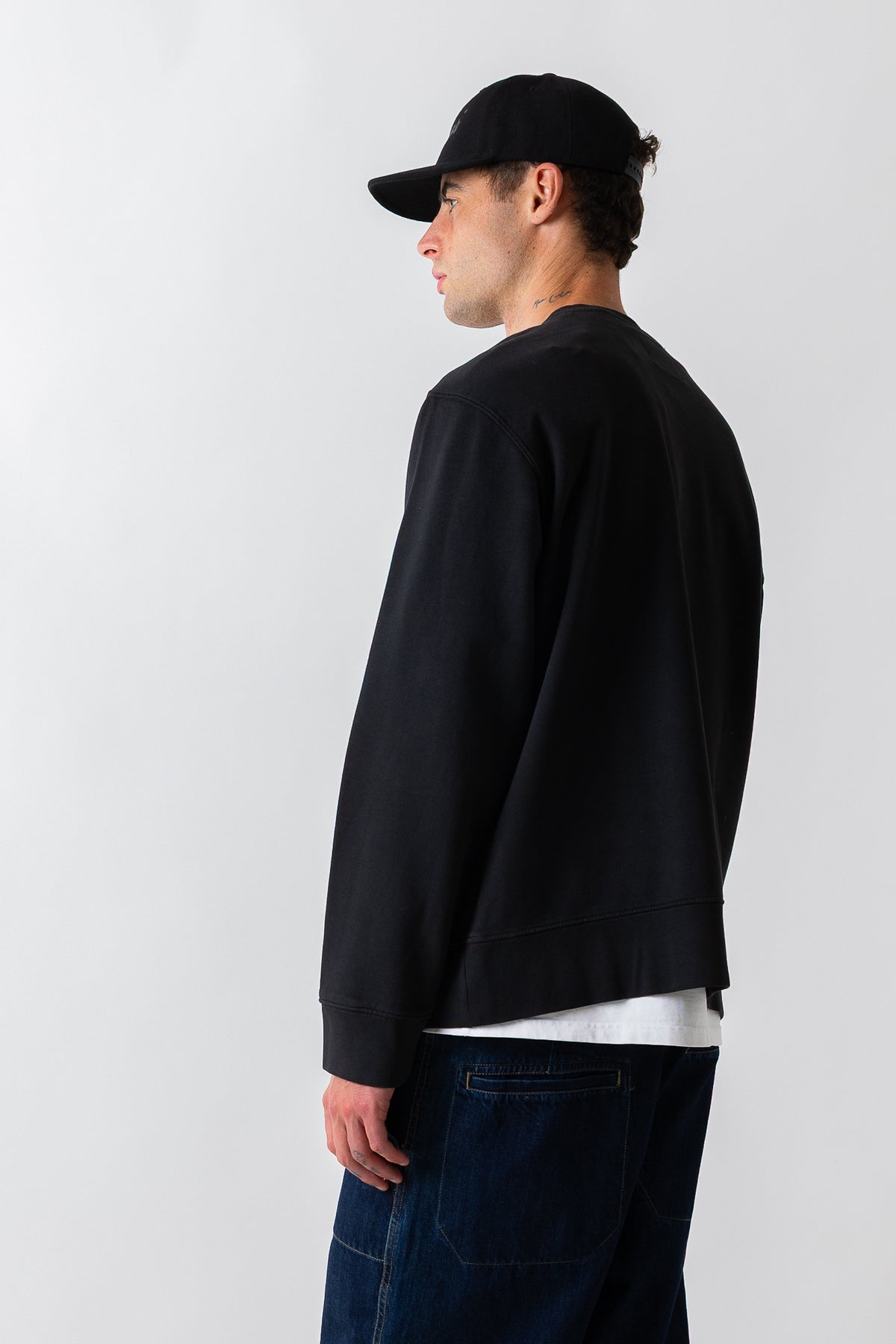 A man in profile wearing a black crewneck sweatshirt, a black baseball cap, and dark blue jeans, photographed against a plain light gray studio background.