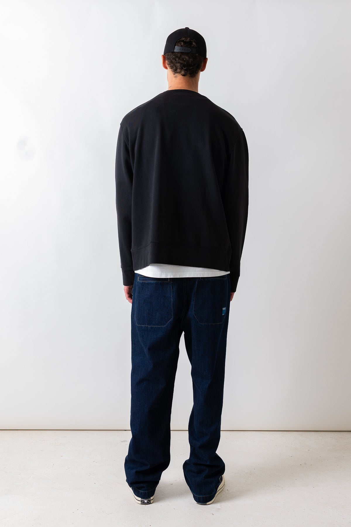 A man viewed from behind wearing a black long-sleeve crewneck sweatshirt, dark blue jeans, a black baseball cap, and white sneakers, standing against a plain white studio background.