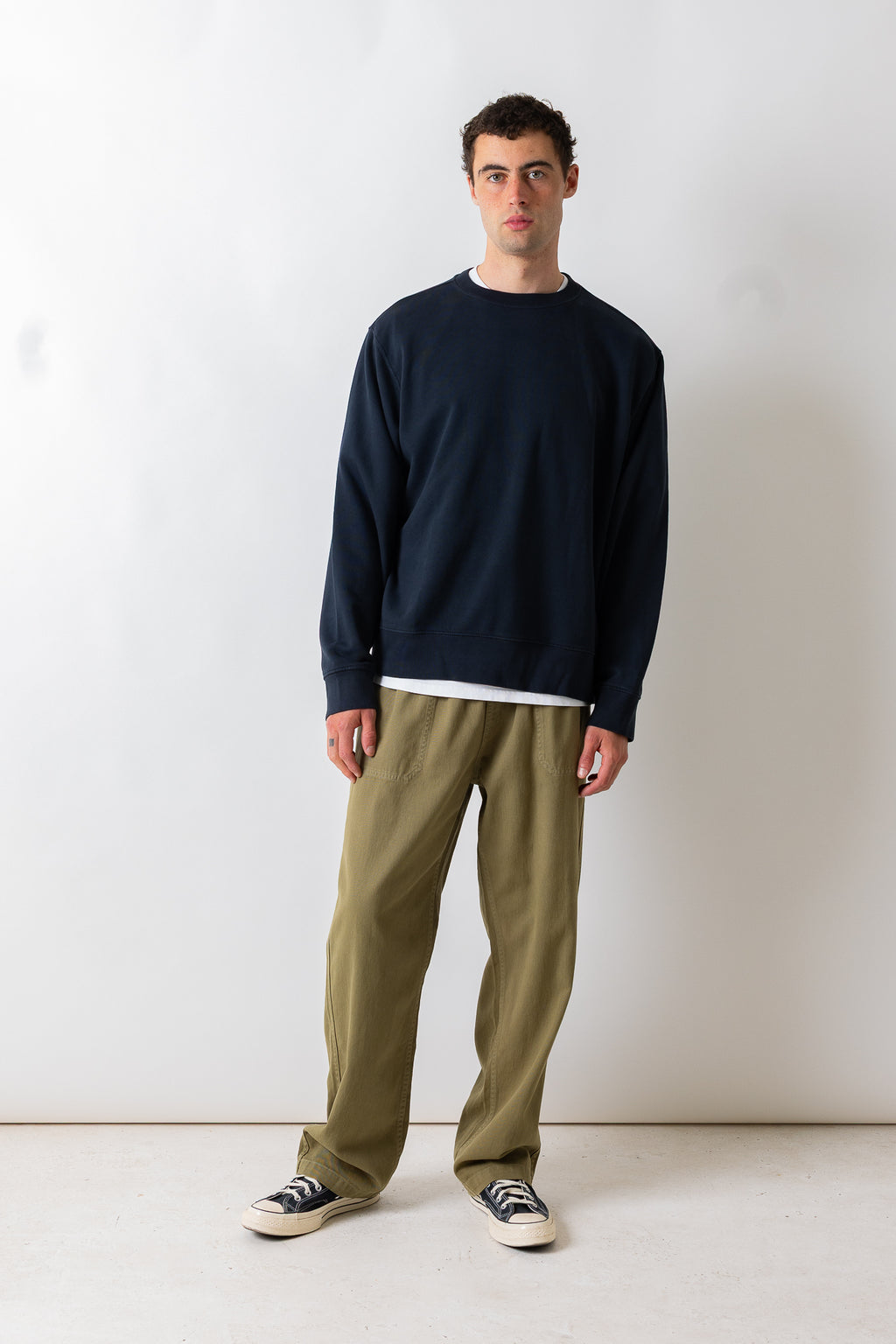 A man wearing a navy blue crewneck sweatshirt, olive green pants, and black-and-white sneakers, standing against a plain off-white studio wall.