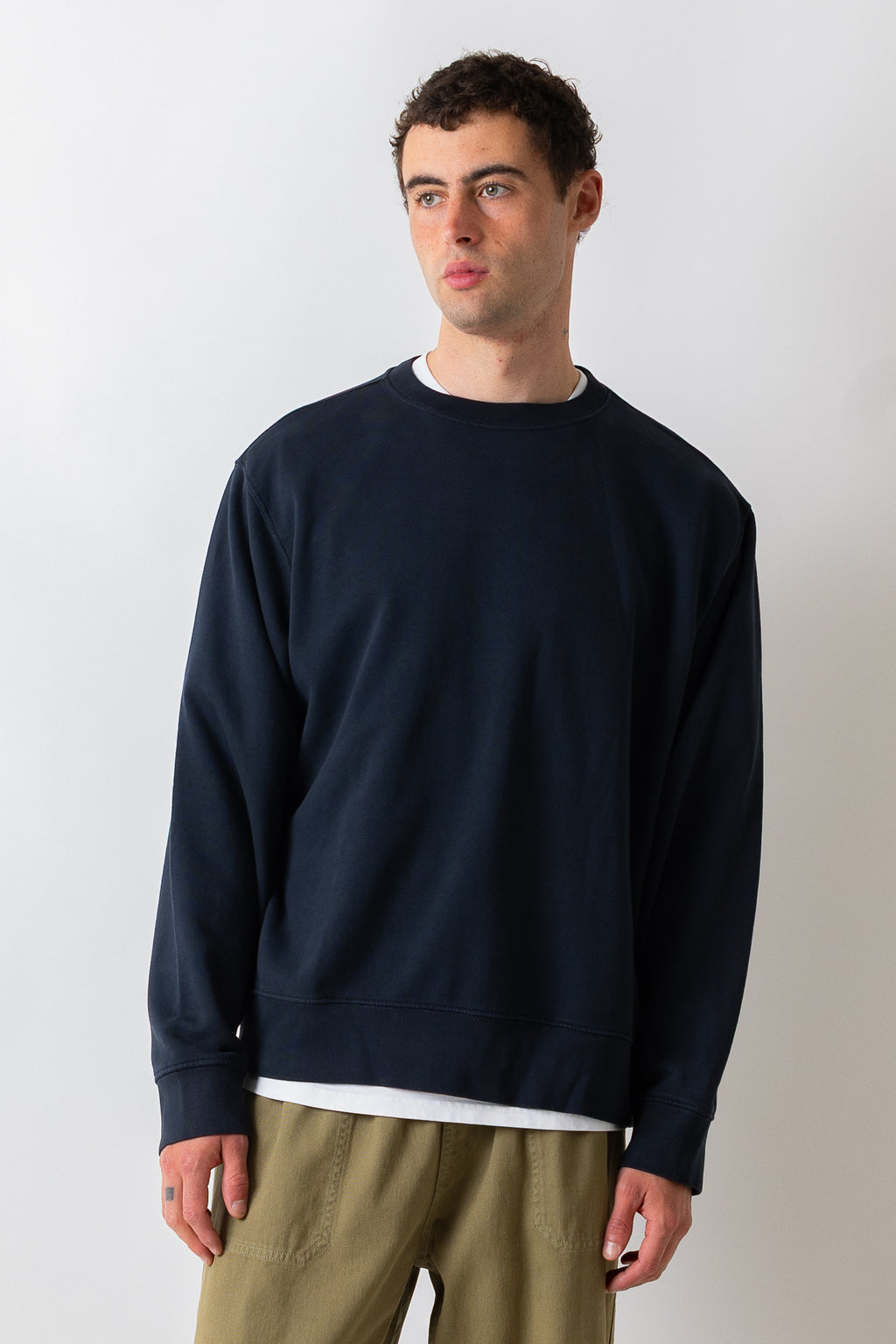 A man wearing a dark navy blue crewneck sweatshirt over a white t-shirt and khaki pants, standing against a plain light gray background.