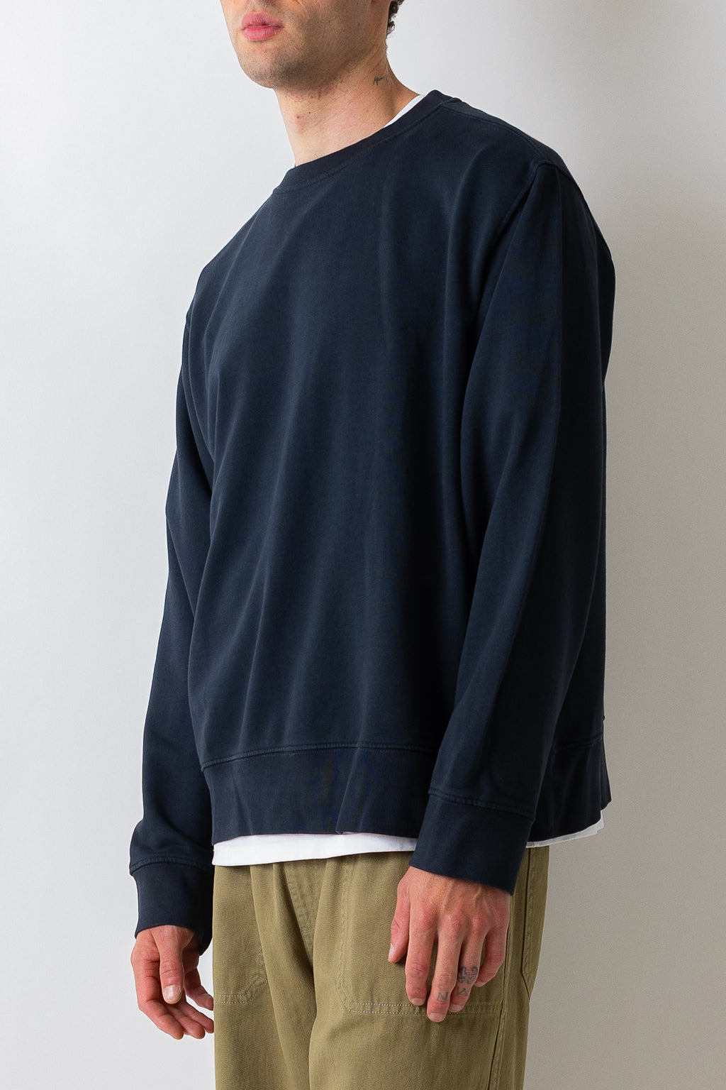 A man wearing a dark navy blue crewneck sweatshirt over a white undershirt and olive green pants, shown from the side against a plain light gray background.