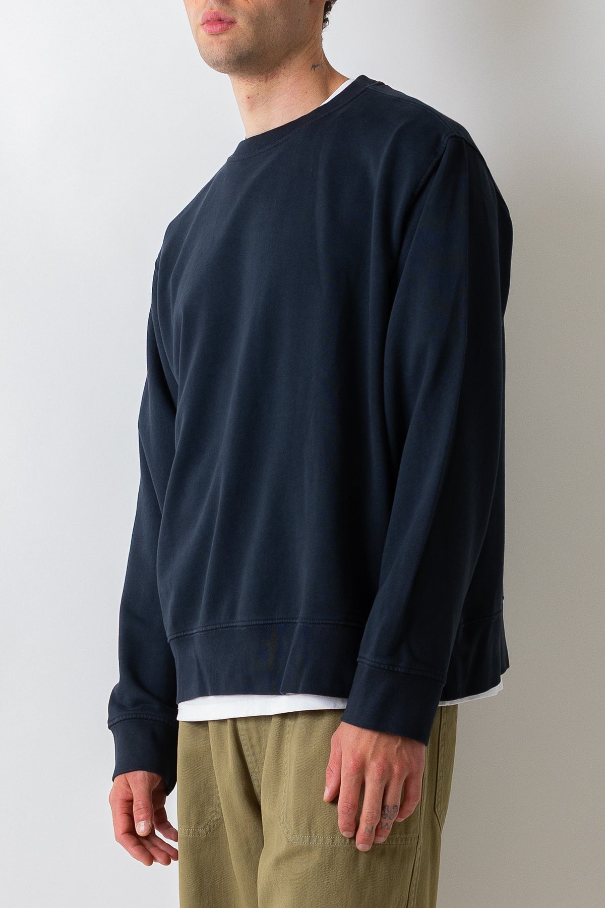 A man wearing a dark navy blue crewneck sweatshirt over a white undershirt and olive green pants, shown from the side against a plain light gray background.