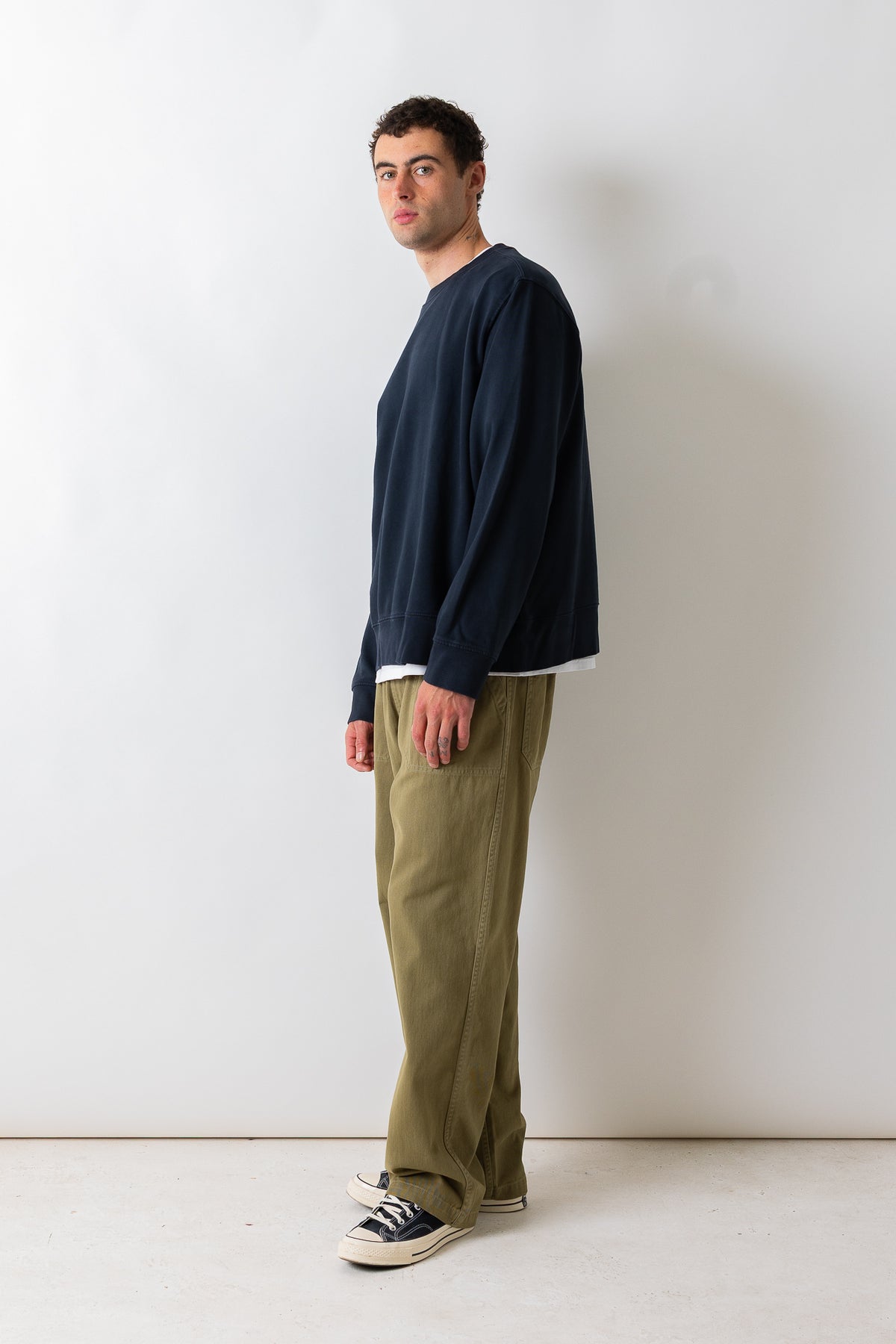 A man stands in profile wearing a dark navy long-sleeve crewneck sweatshirt, olive green pants, and black-and-white sneakers against a plain off-white studio wall.
