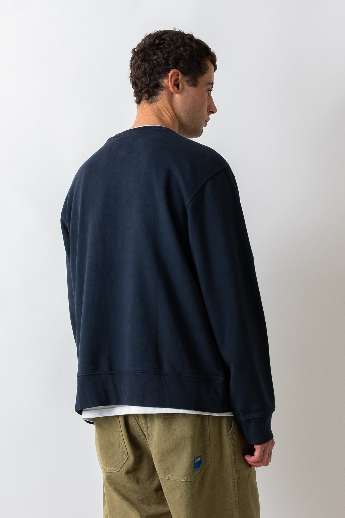 A man shown from the back wearing a dark navy crewneck sweatshirt, olive green pants, and a white t-shirt underneath, standing against a plain light gray background.