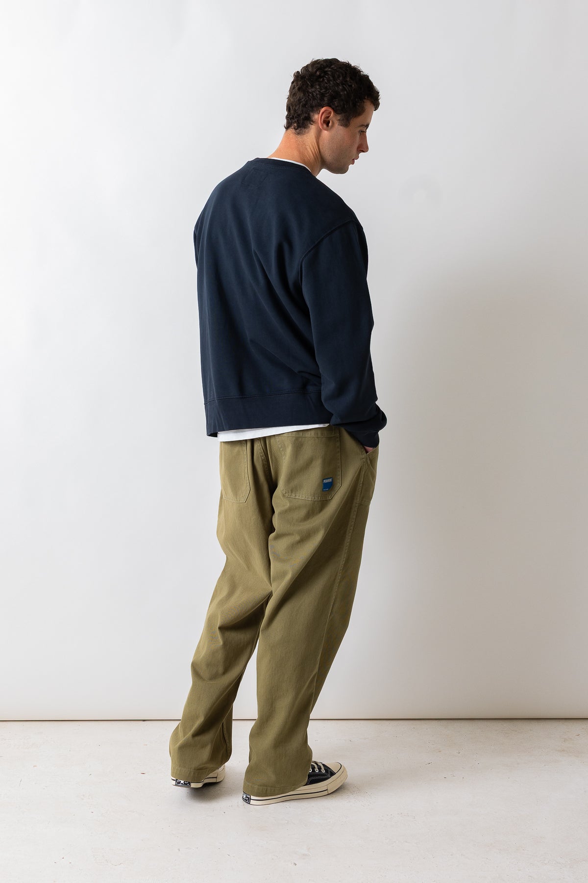 A man wearing a navy blue crewneck sweatshirt, olive green relaxed-fit pants, and black canvas sneakers, standing in profile against a plain off-white studio wall.