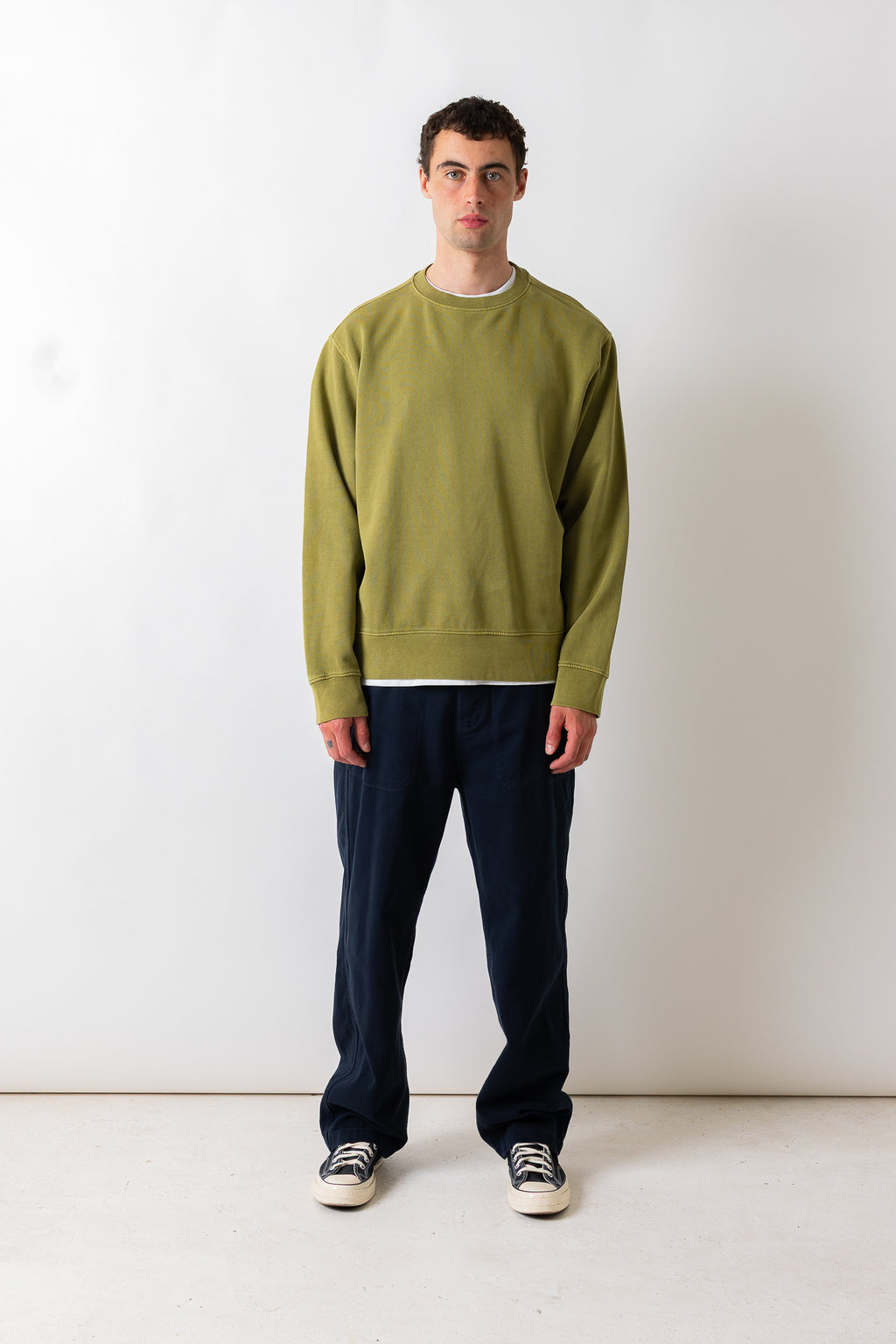 A man wearing a muted olive green crewneck sweatshirt and dark navy pants, standing against a plain light gray studio background.