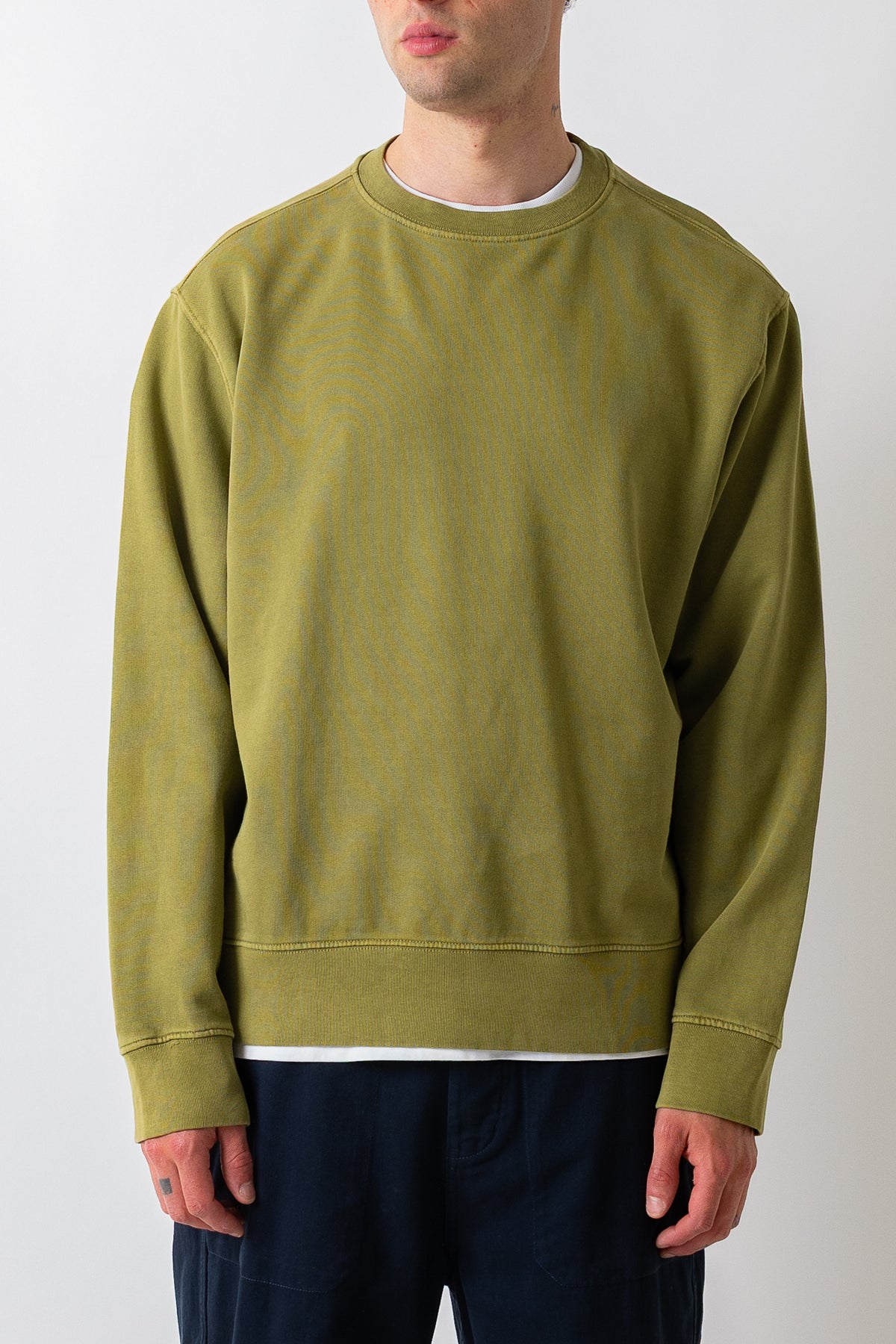 A man wearing a muted olive green crewneck sweatshirt and dark navy pants, standing against a plain light gray studio background.