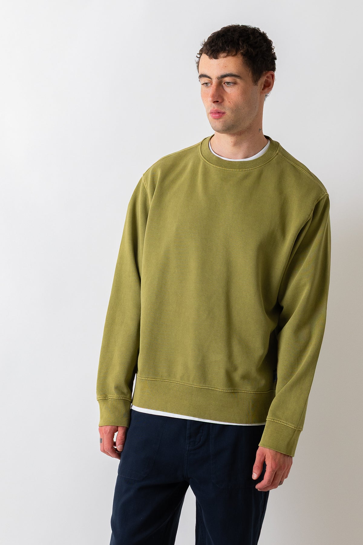 A man wearing a muted olive green crewneck sweatshirt and dark navy pants, standing against a plain light gray studio background.