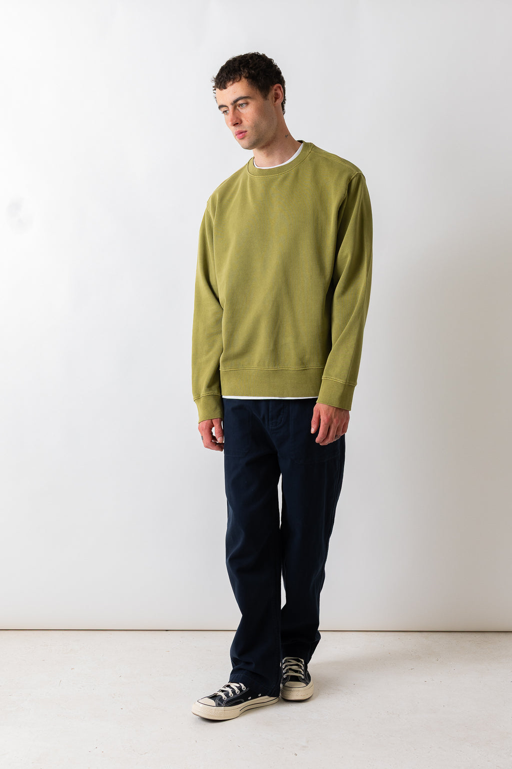 A man wearing an olive green crewneck sweatshirt, dark navy pants, and black canvas sneakers, standing against a plain white studio background.