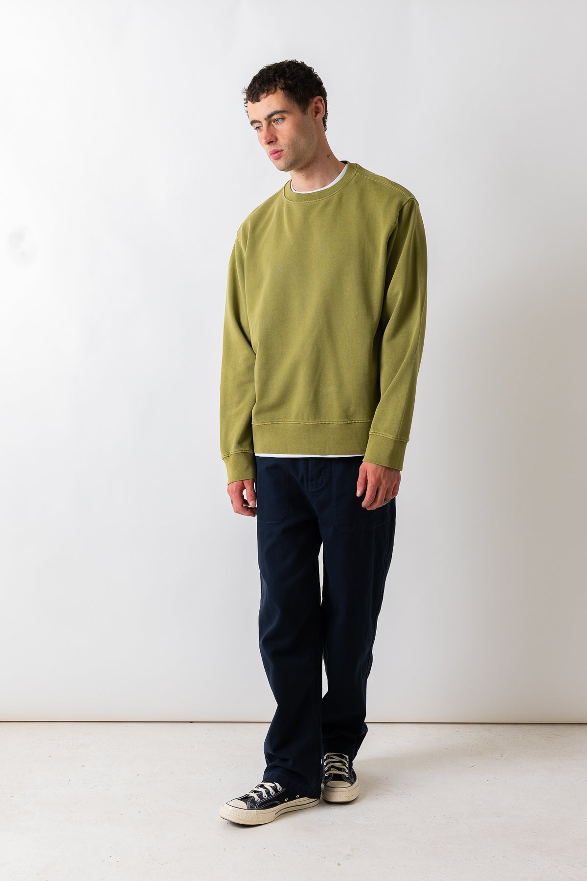 A man wearing an olive green crewneck sweatshirt, dark navy pants, and black canvas sneakers, standing against a plain white studio background.