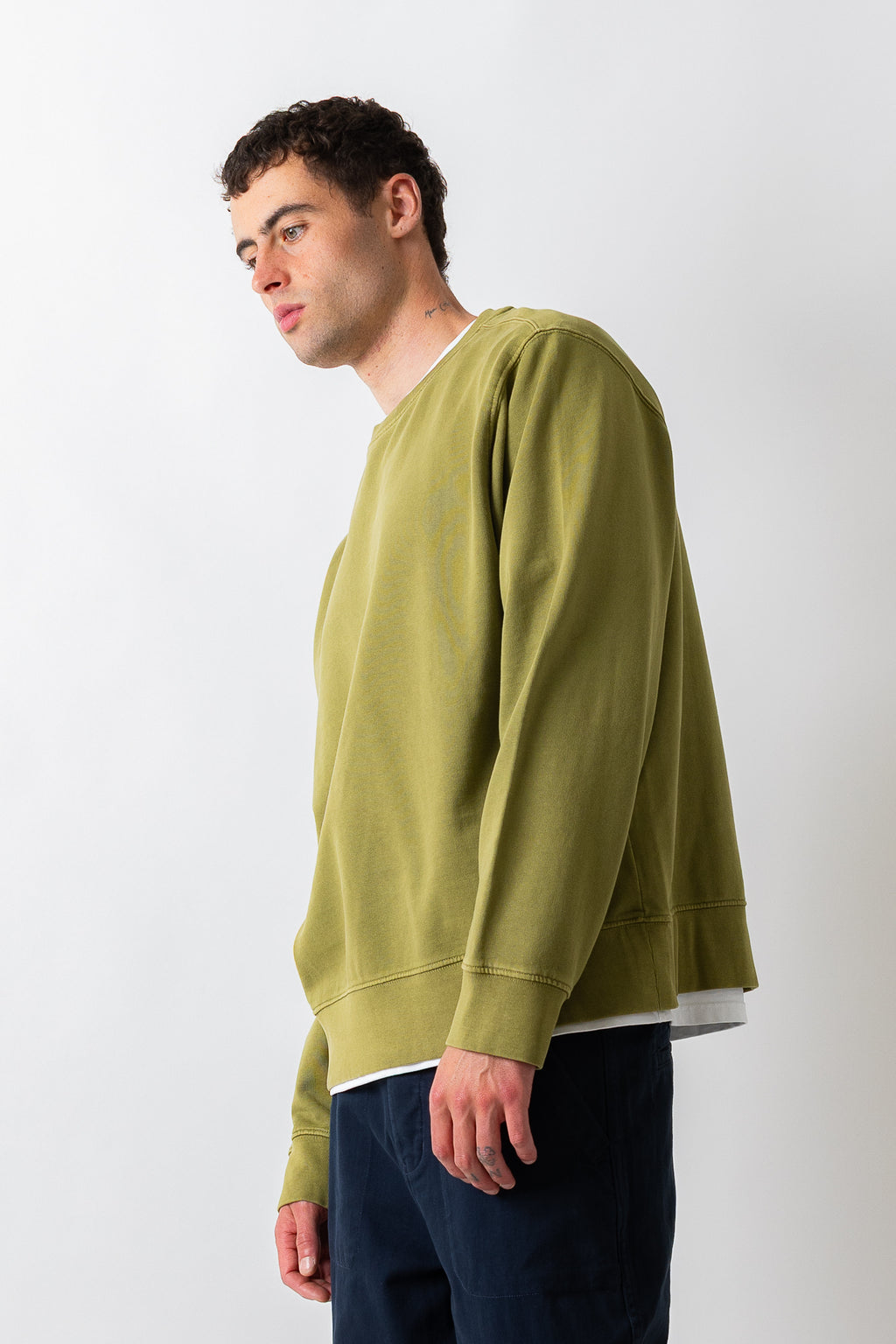 A man wearing a muted olive green crewneck sweatshirt and dark navy pants, standing against a plain light gray studio background.