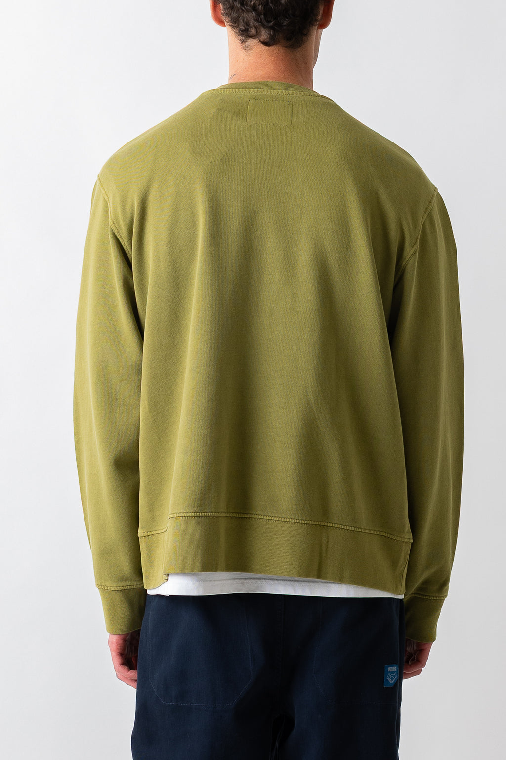A man viewed from behind wearing an olive green crewneck sweatshirt and dark navy pants against a plain light gray background.