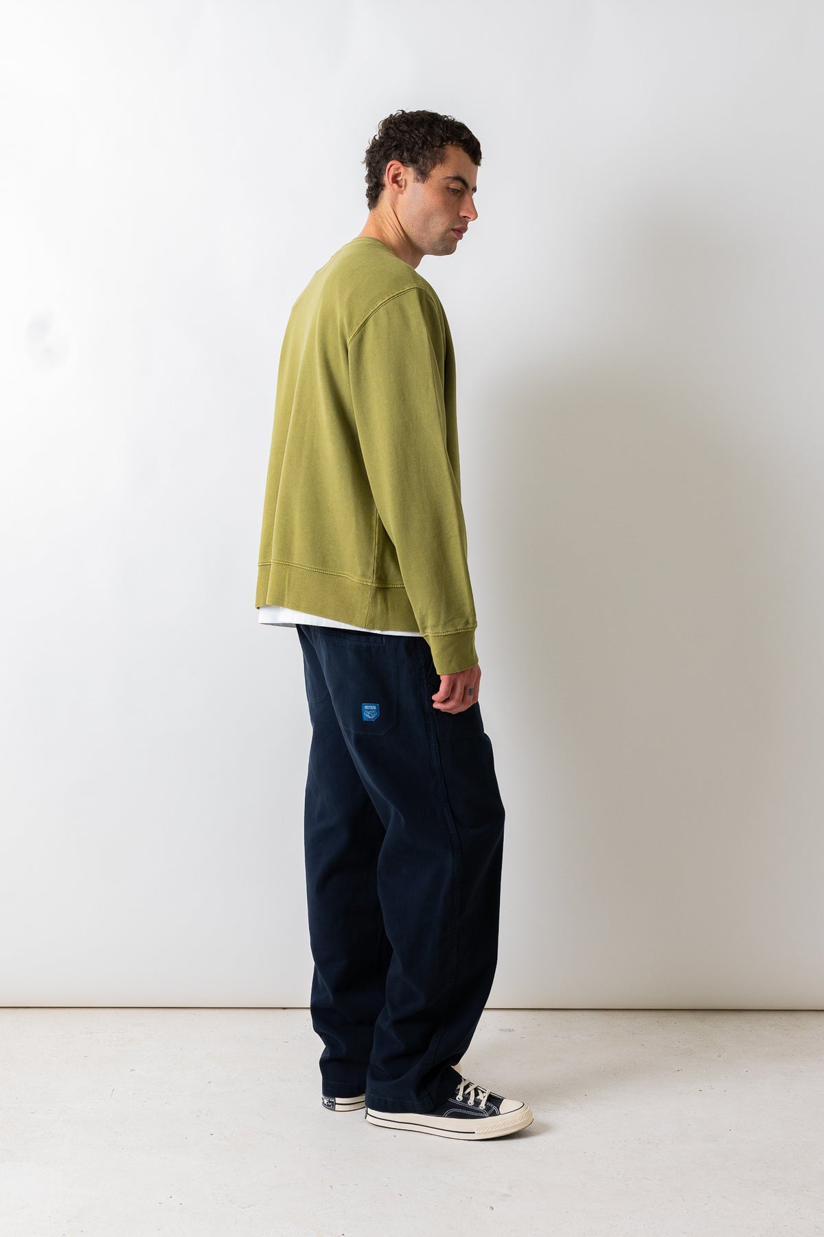 A man wearing a sage green crewneck sweatshirt, dark navy wide-leg pants, and black canvas sneakers, standing in profile against a plain off-white studio background.