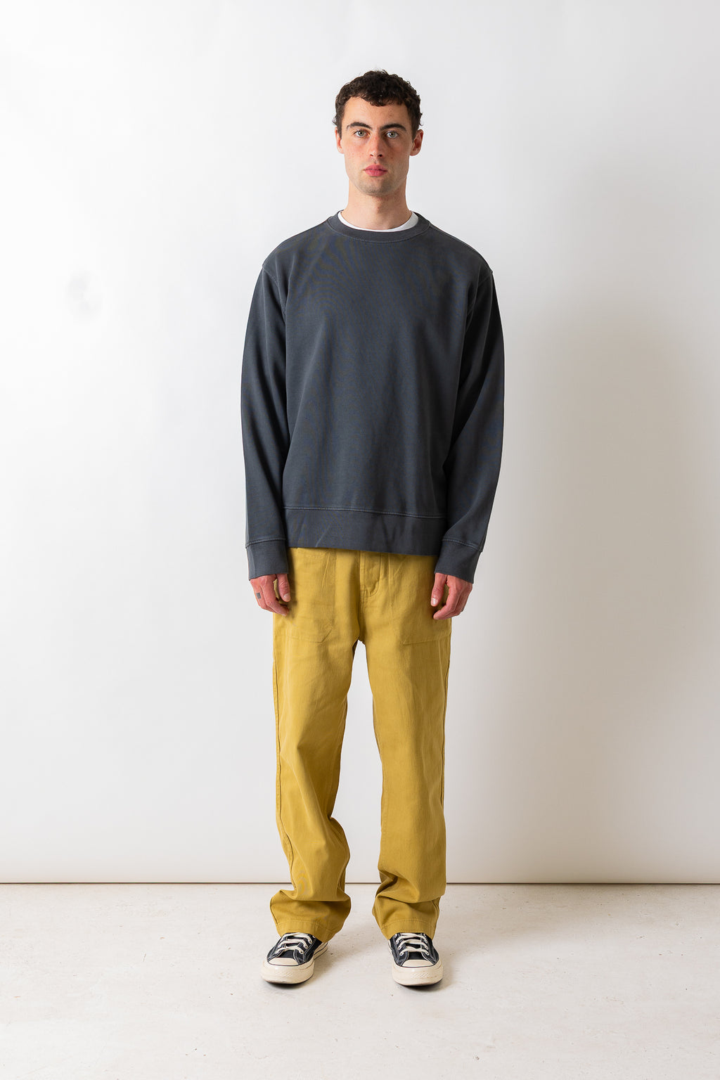 A man stands against a plain off-white wall wearing a dark charcoal gray crewneck sweatshirt, mustard yellow pants, and black and white sneakers.