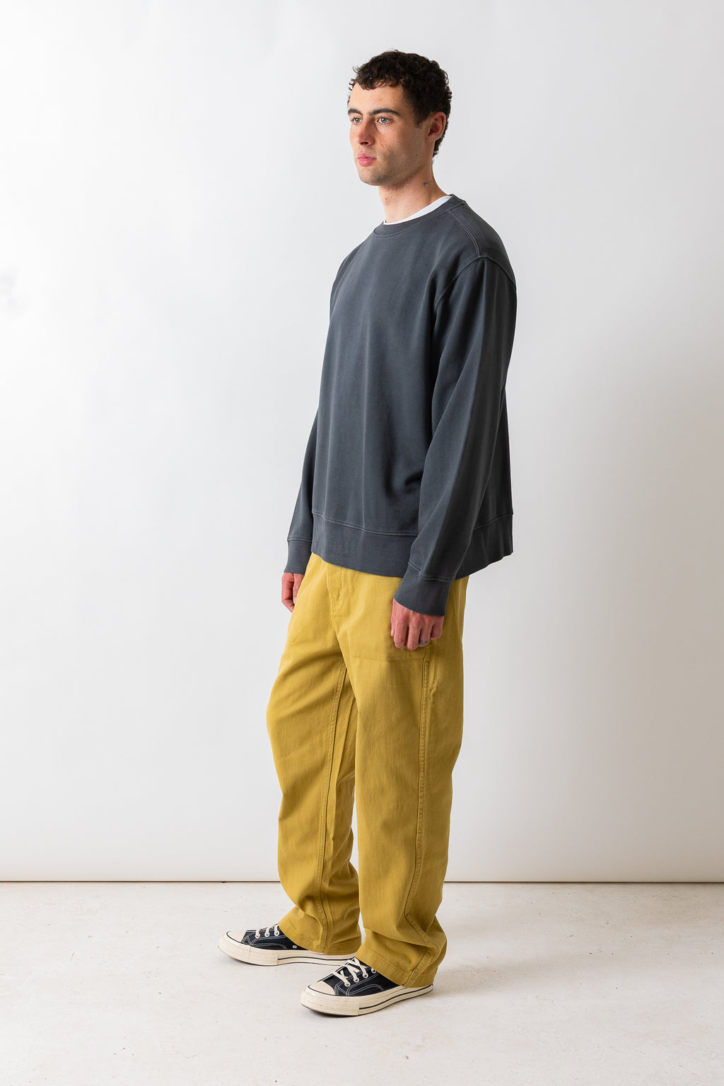 A man wearing a dark charcoal gray crewneck sweatshirt, mustard yellow corduroy pants, and black canvas sneakers, standing in profile against a plain white studio background.