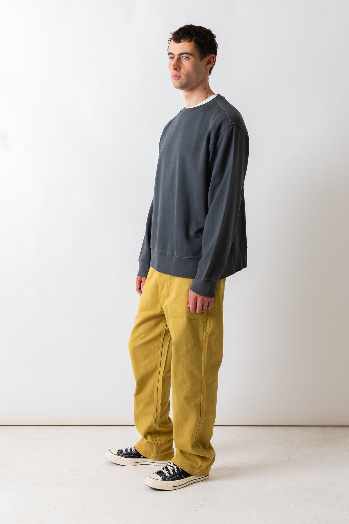 A man wearing a dark charcoal gray crewneck sweatshirt, mustard yellow corduroy pants, and black canvas sneakers, standing in profile against a plain white studio background.