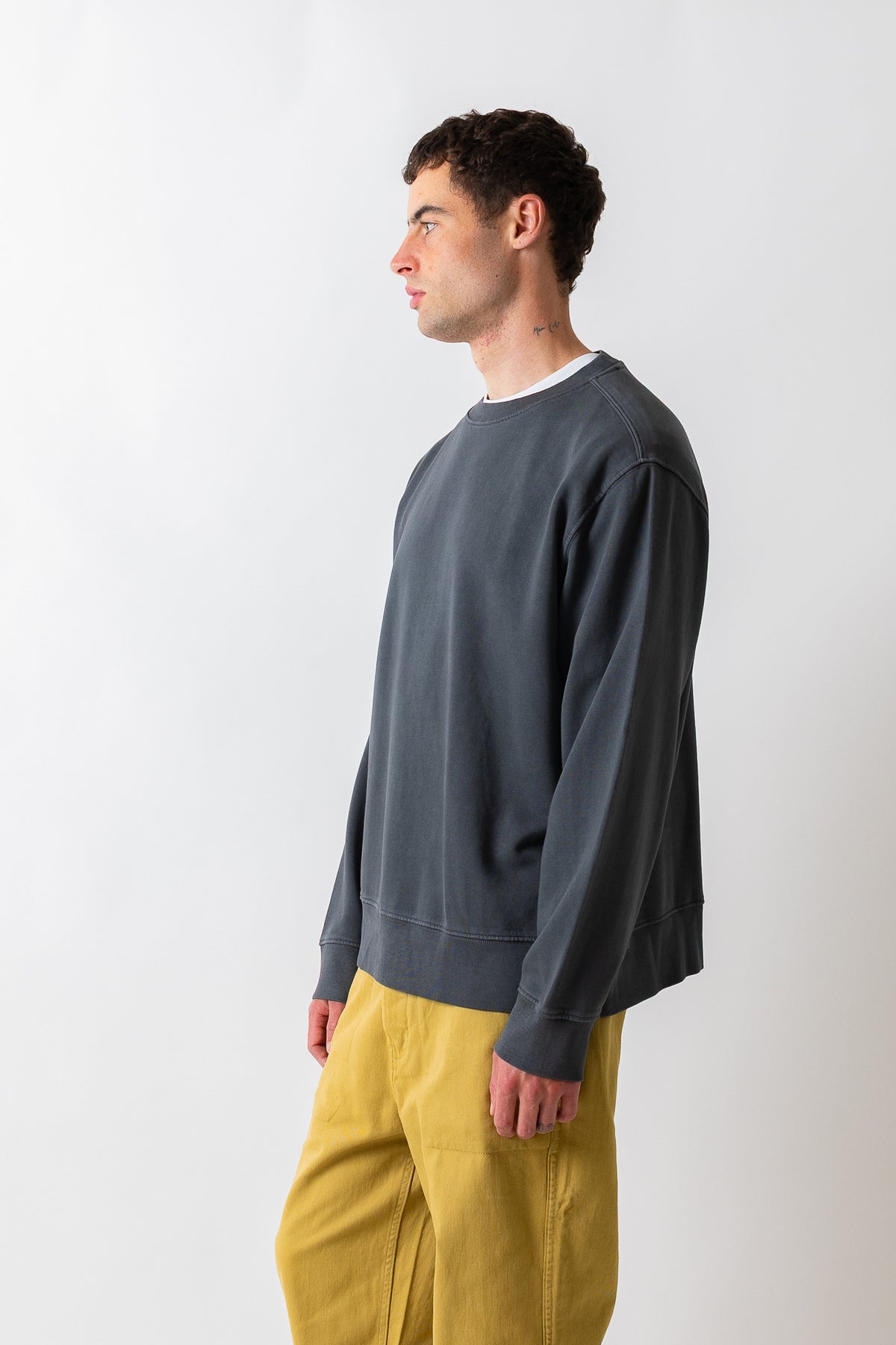A man in profile wearing a dark charcoal gray crewneck sweatshirt and mustard yellow pants, standing against a plain light gray studio background.