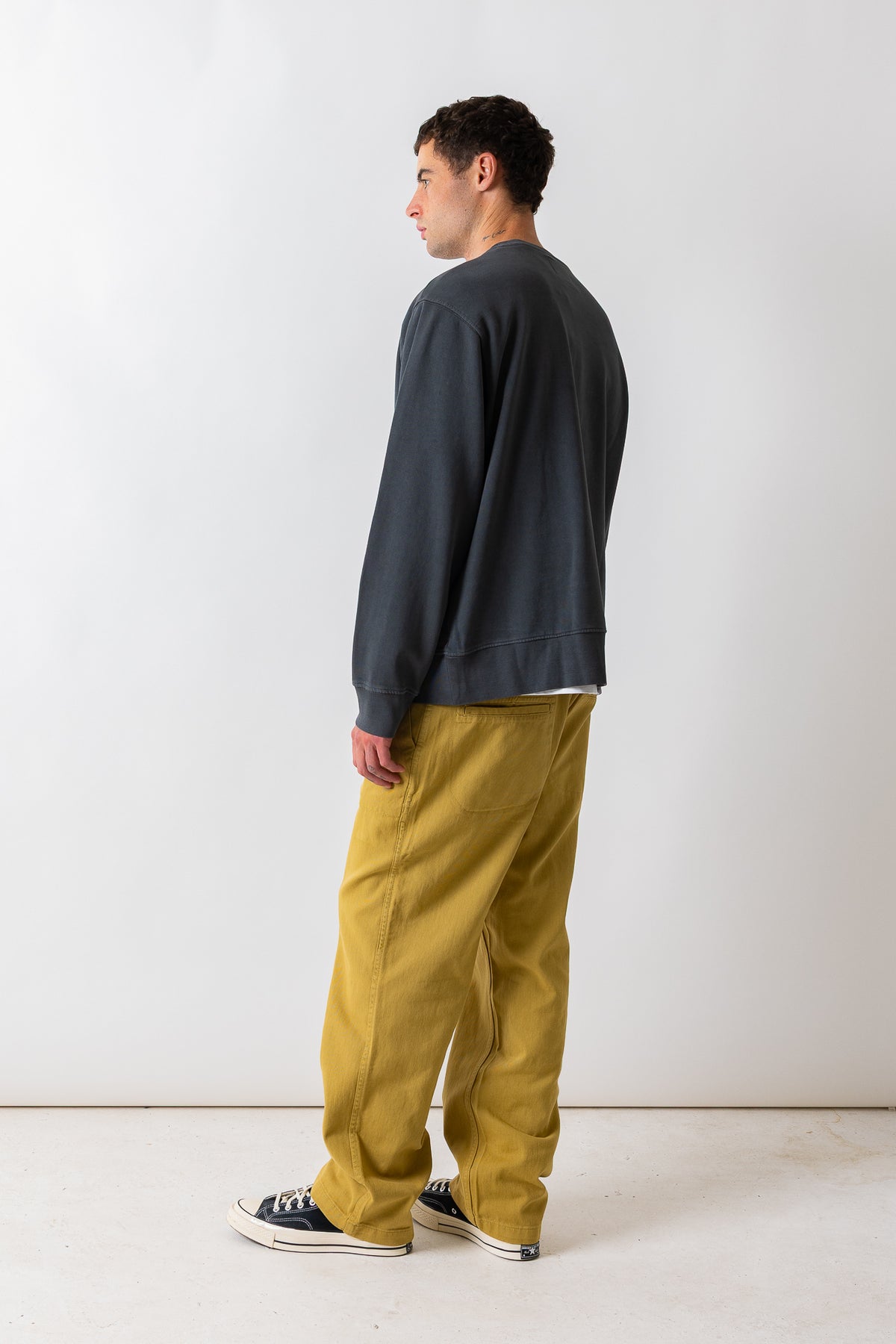 A man standing in profile wearing a dark charcoal crewneck sweatshirt, mustard yellow wide-leg pants, and black canvas sneakers against a plain white studio background.