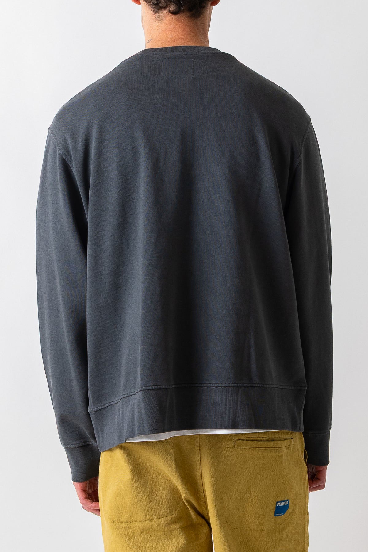 A man viewed from behind wearing a dark charcoal gray crewneck sweatshirt and mustard yellow pants against a light gray studio background.
