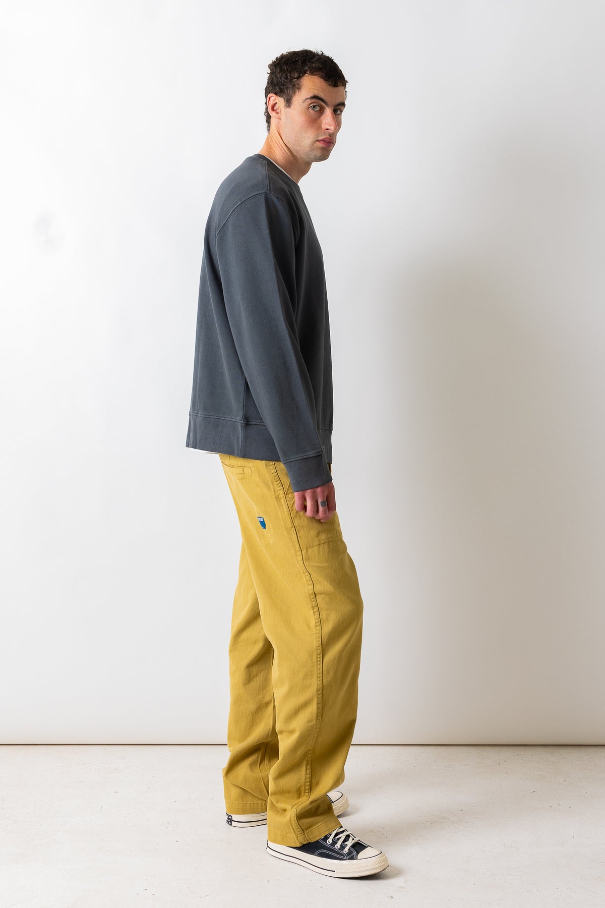A man wearing a dark gray crewneck sweatshirt, mustard yellow wide-leg pants, and navy blue canvas sneakers, standing in profile against a plain off-white wall.