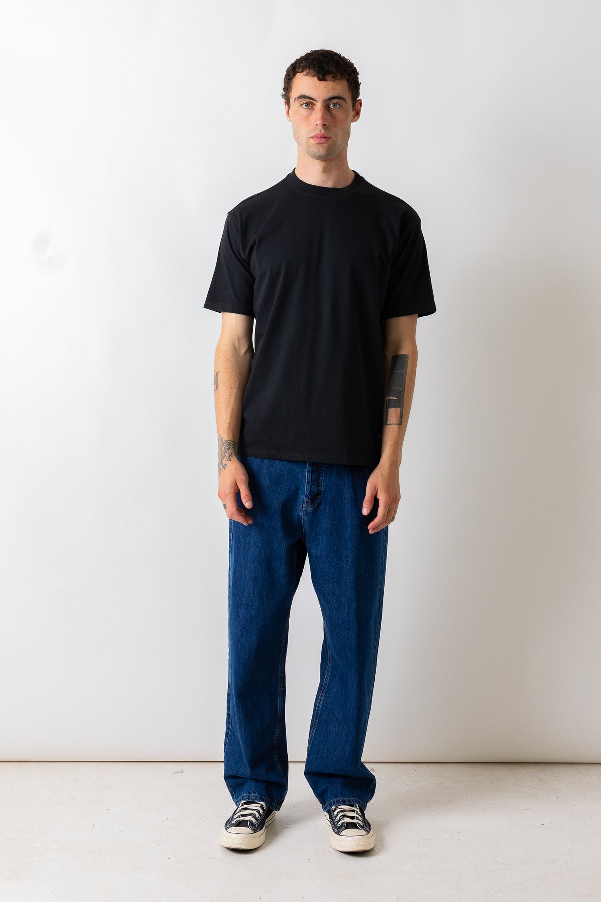 A man wearing a black short-sleeve crewneck t-shirt, loose blue jeans, and black and white sneakers, standing against a plain off-white wall.
