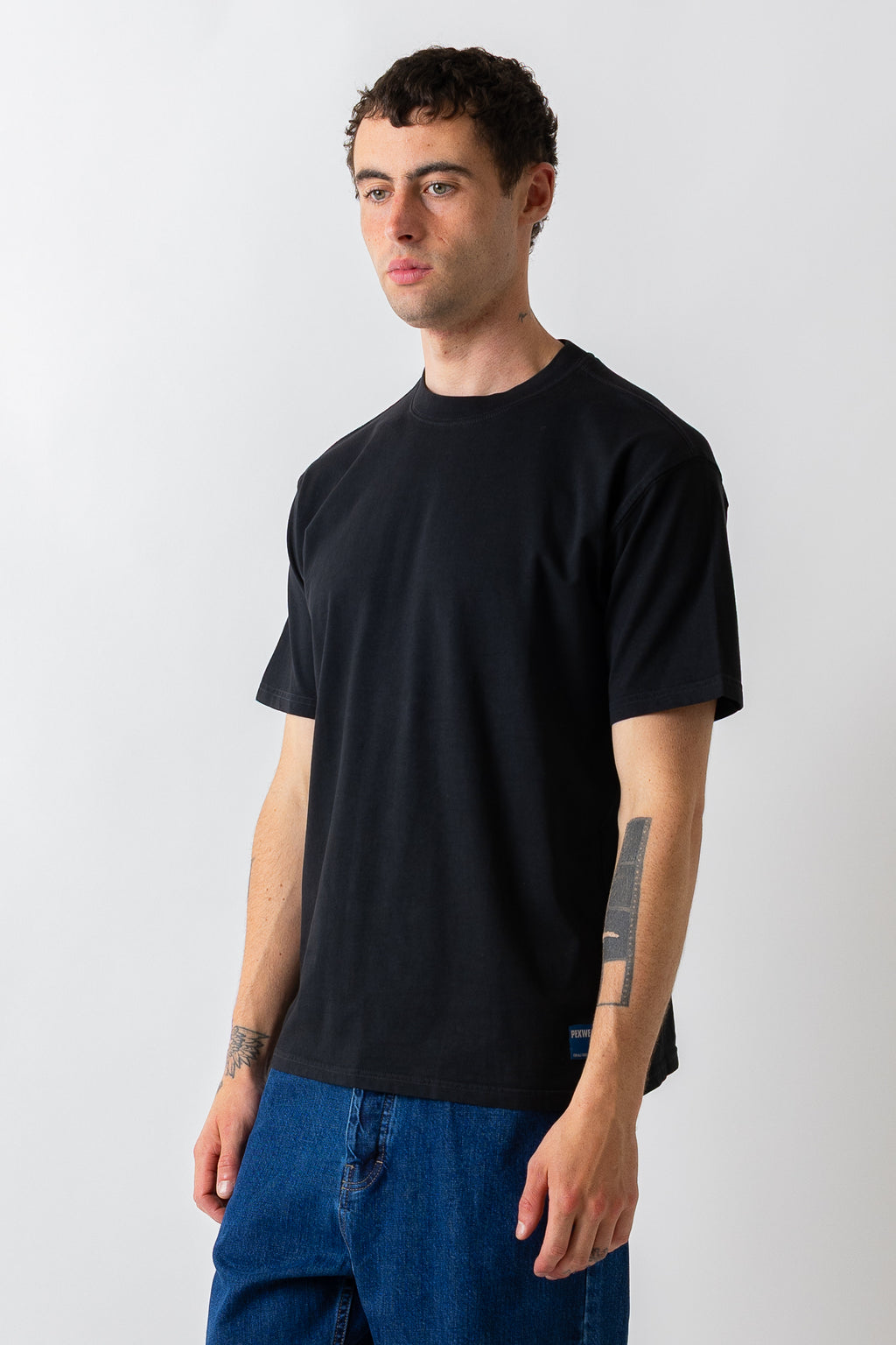 A man wearing a plain black short-sleeve crewneck t-shirt and blue jeans, standing against a light gray studio background.