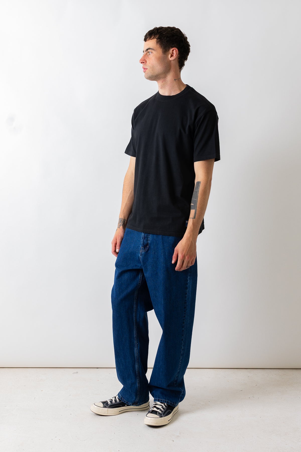 A man wearing a black crewneck t-shirt, loose-fit dark blue jeans, and black canvas sneakers, standing in profile against a plain white studio background.