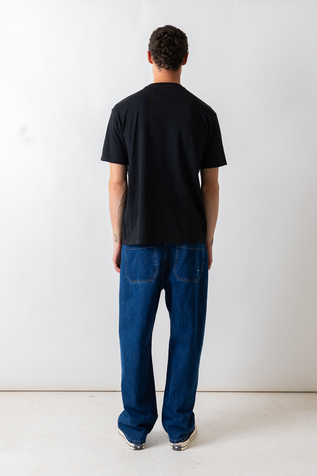 A man viewed from behind wearing a black short-sleeve t-shirt and loose-fit blue jeans, standing against a plain white studio wall.