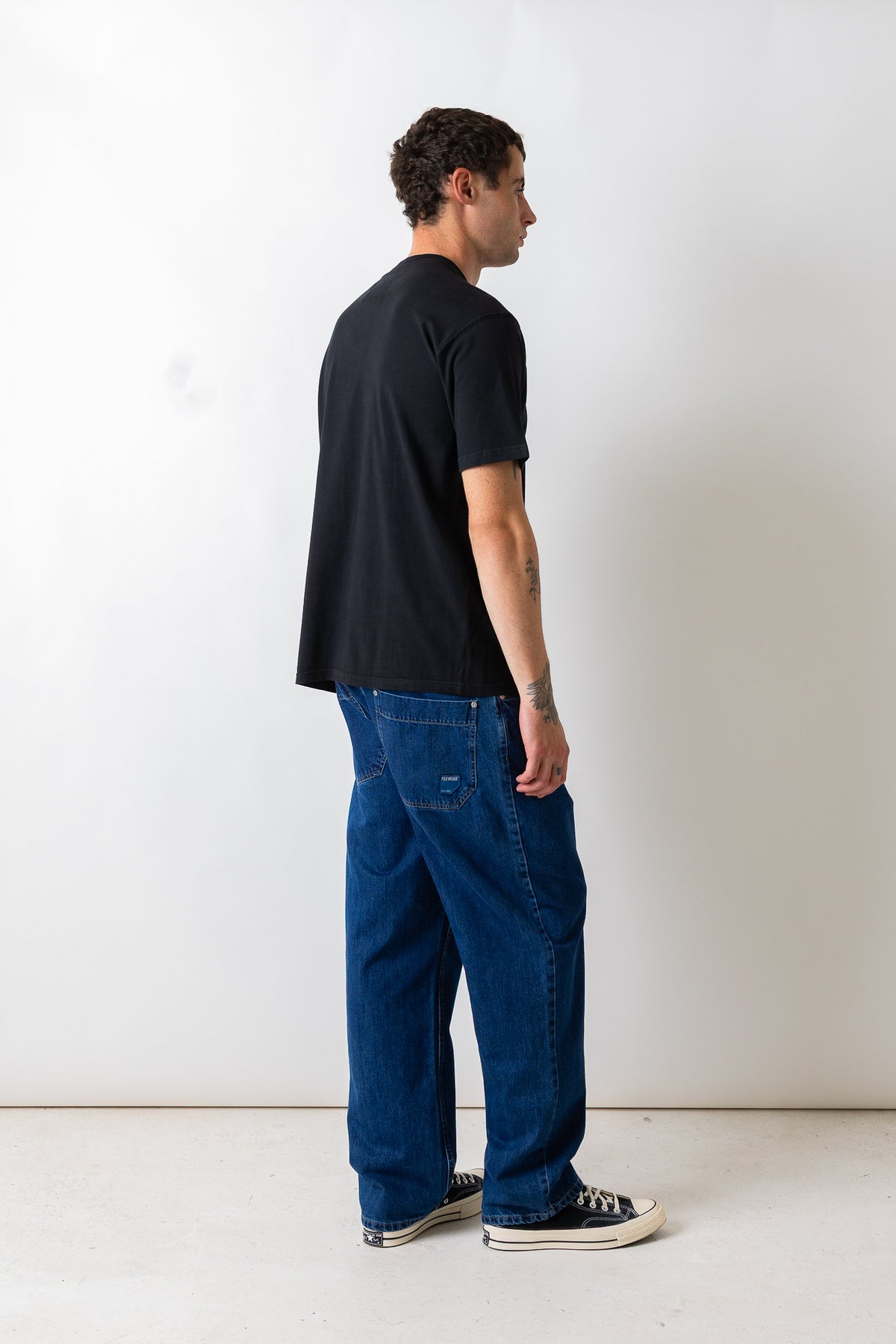 A man standing in profile wearing a black t-shirt, blue denim jeans, and black canvas sneakers against a plain white studio background.