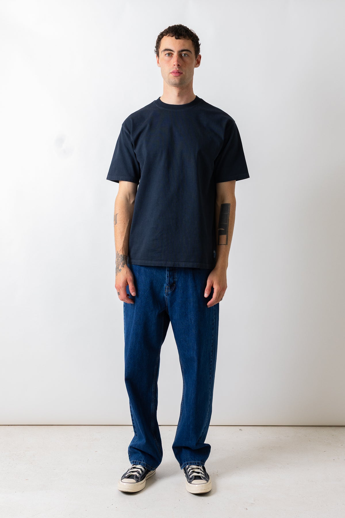 A man stands against a plain white studio wall wearing a dark navy blue short-sleeve crewneck t-shirt, dark blue jeans, and black and white sneakers.