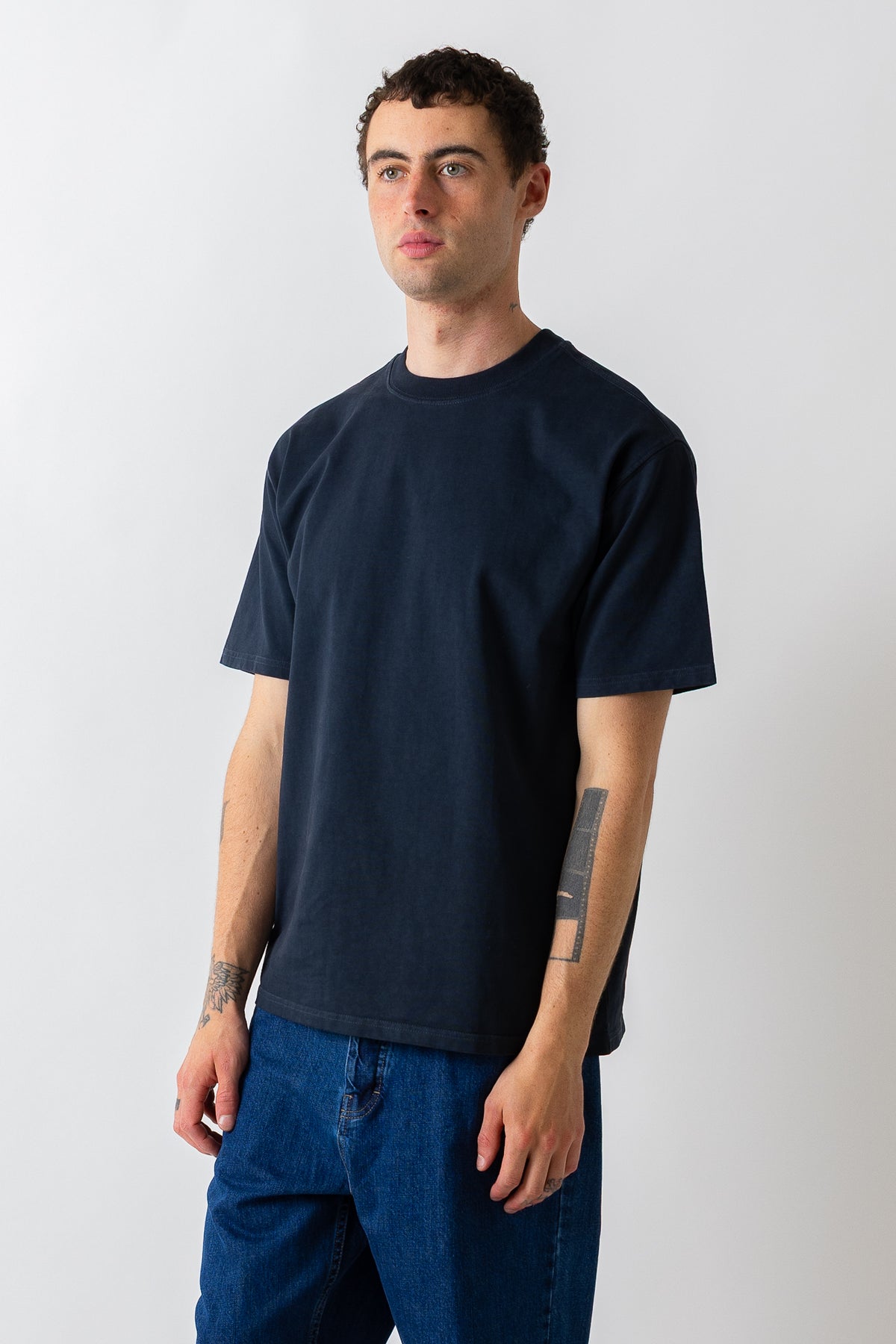 A man wearing a dark navy blue crewneck t-shirt and blue jeans, standing against a light gray studio background.