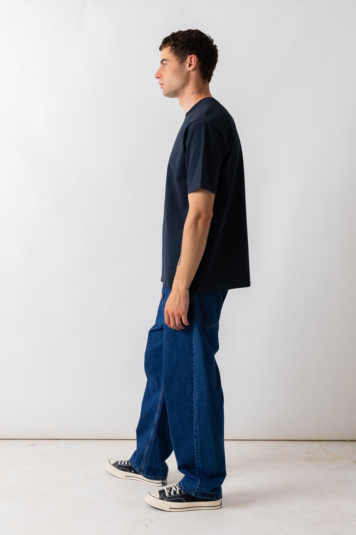 A man in profile wearing a dark navy blue t-shirt, loose-fit blue jeans, and black canvas sneakers, standing against a plain light gray studio wall.