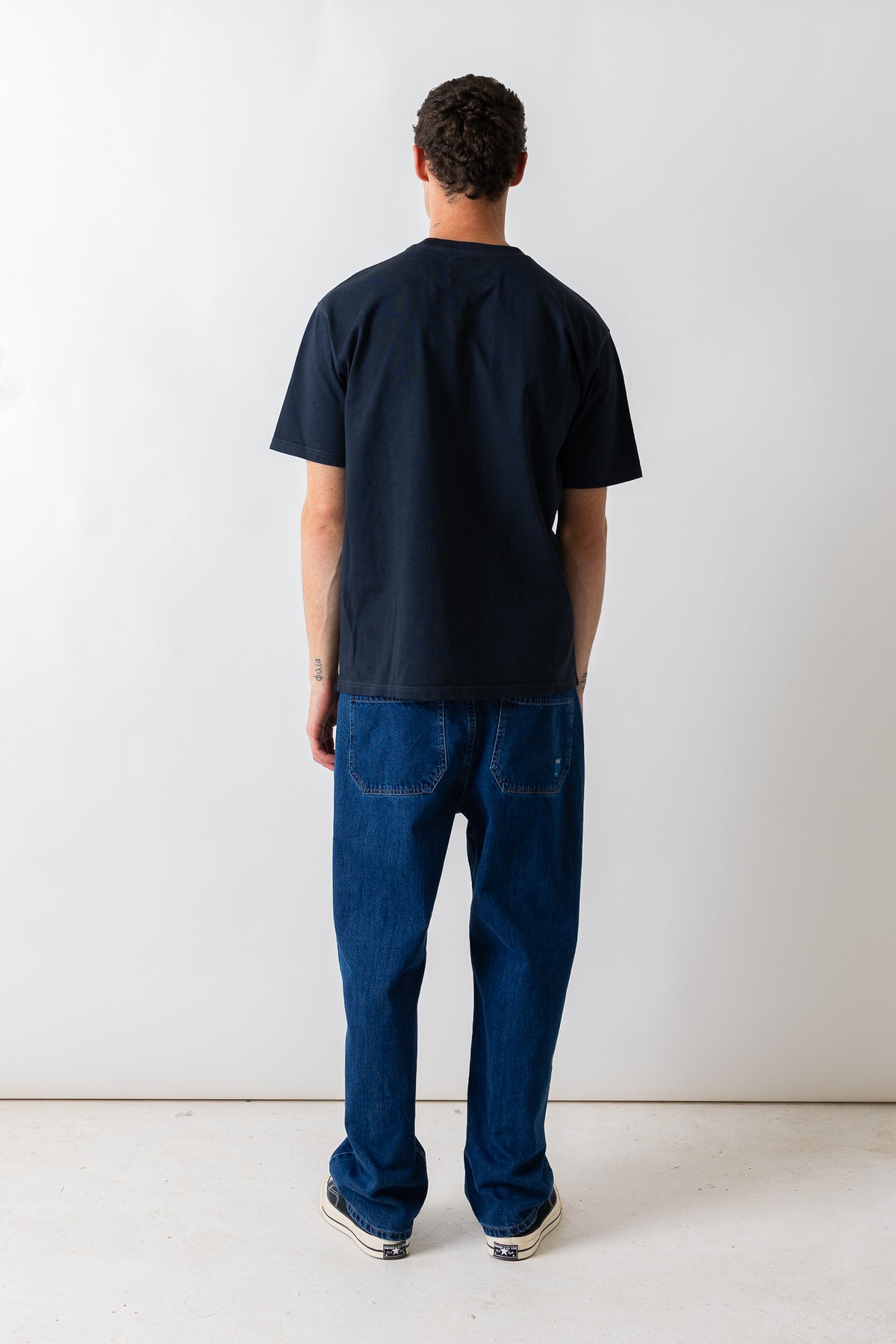 A man viewed from behind wearing a dark navy blue t-shirt and blue jeans, standing against a plain white studio wall.