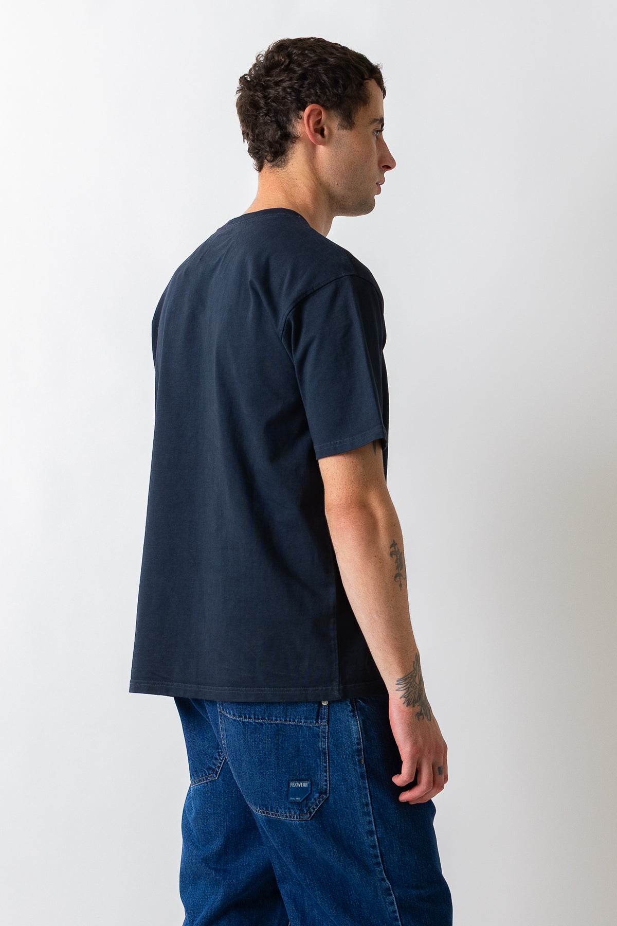 A man shown from the back and side profile wearing a dark navy blue short-sleeve t-shirt and blue jeans against a plain light gray background.