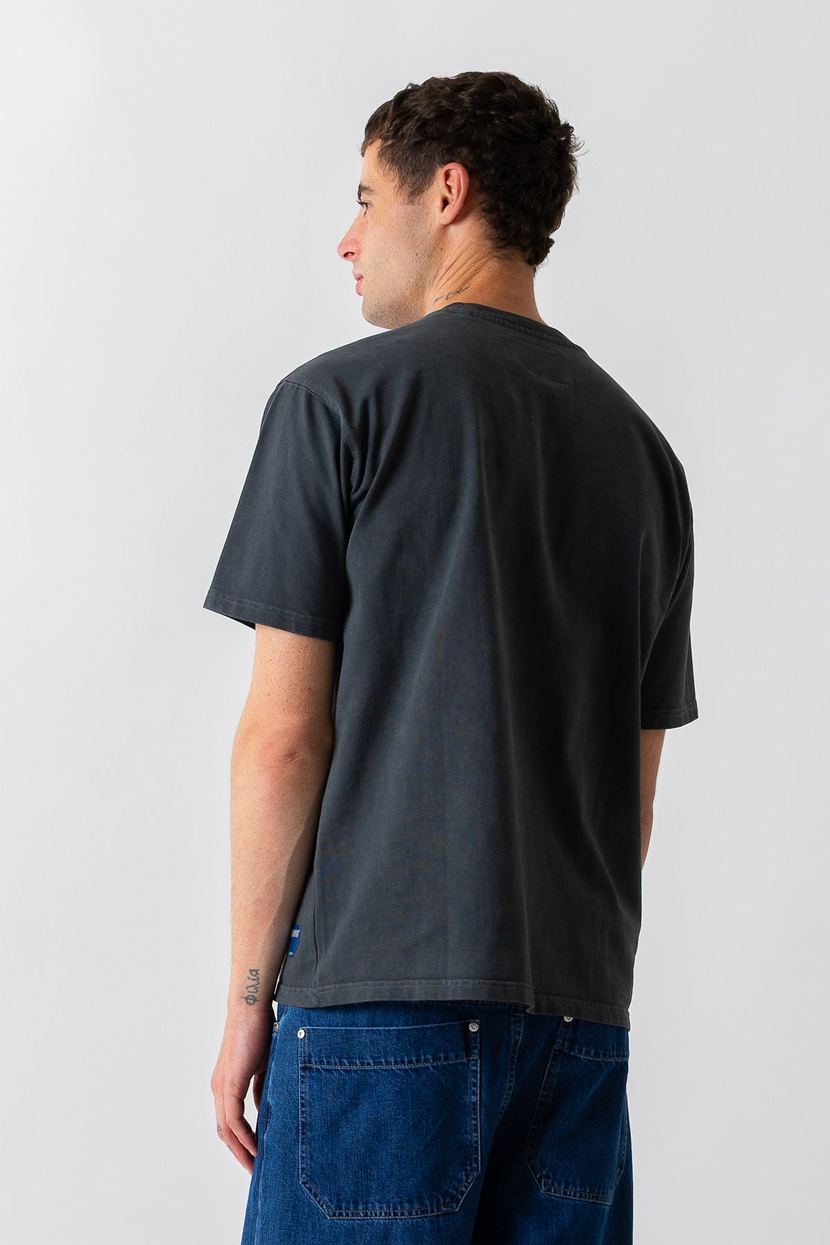 A man wearing a faded black crewneck t-shirt and blue jeans, shown from a back-three-quarter view against a light gray studio background.