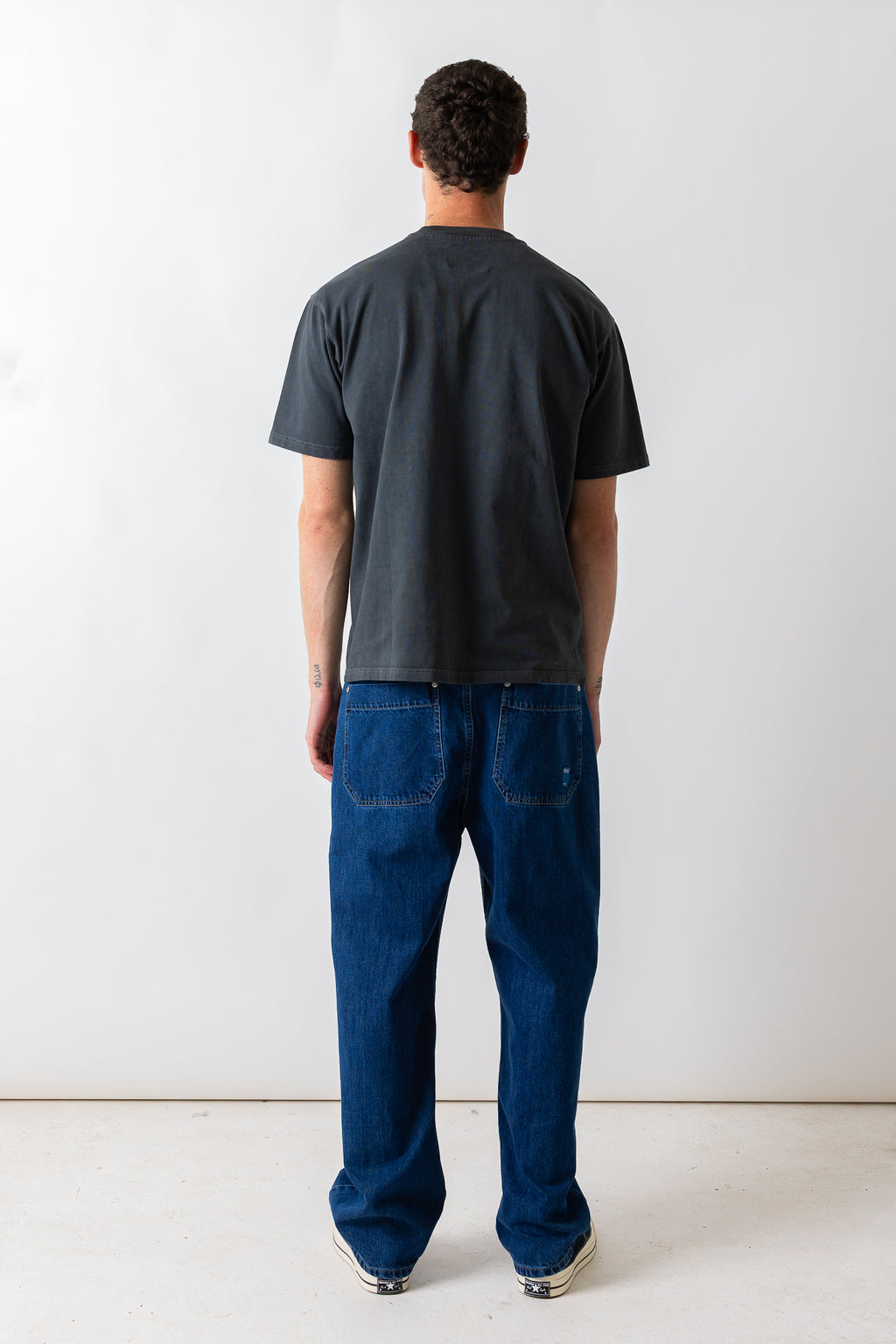 A man viewed from behind wearing a dark charcoal gray t-shirt and blue jeans, standing against a plain white studio wall.