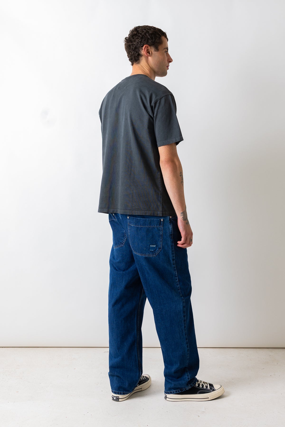 A man wearing a faded black t-shirt and blue jeans, photographed from behind and in profile against a plain white studio background.