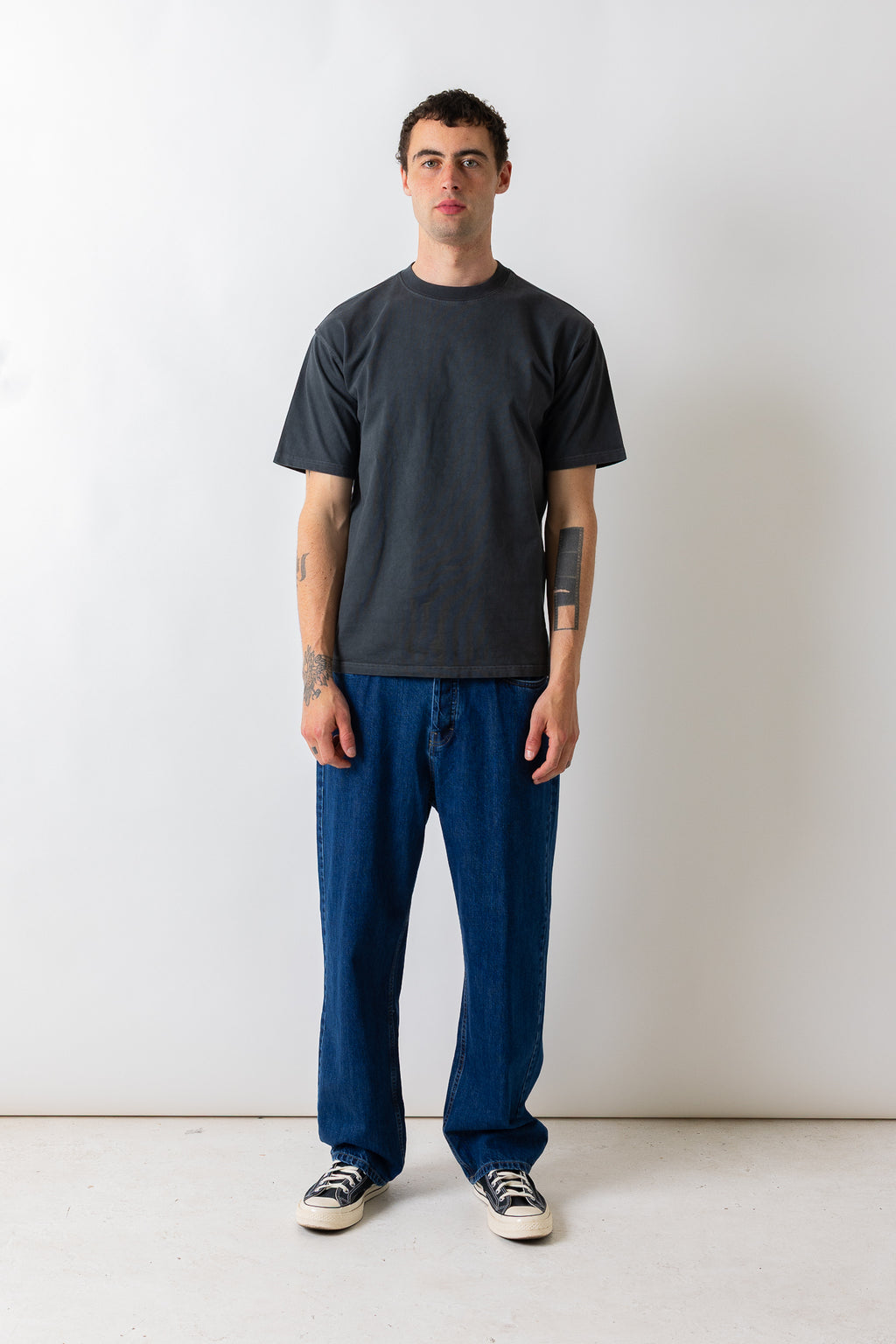 A man wearing a dark charcoal gray crewneck t-shirt, blue jeans, and black and white sneakers, standing against a plain white studio background.